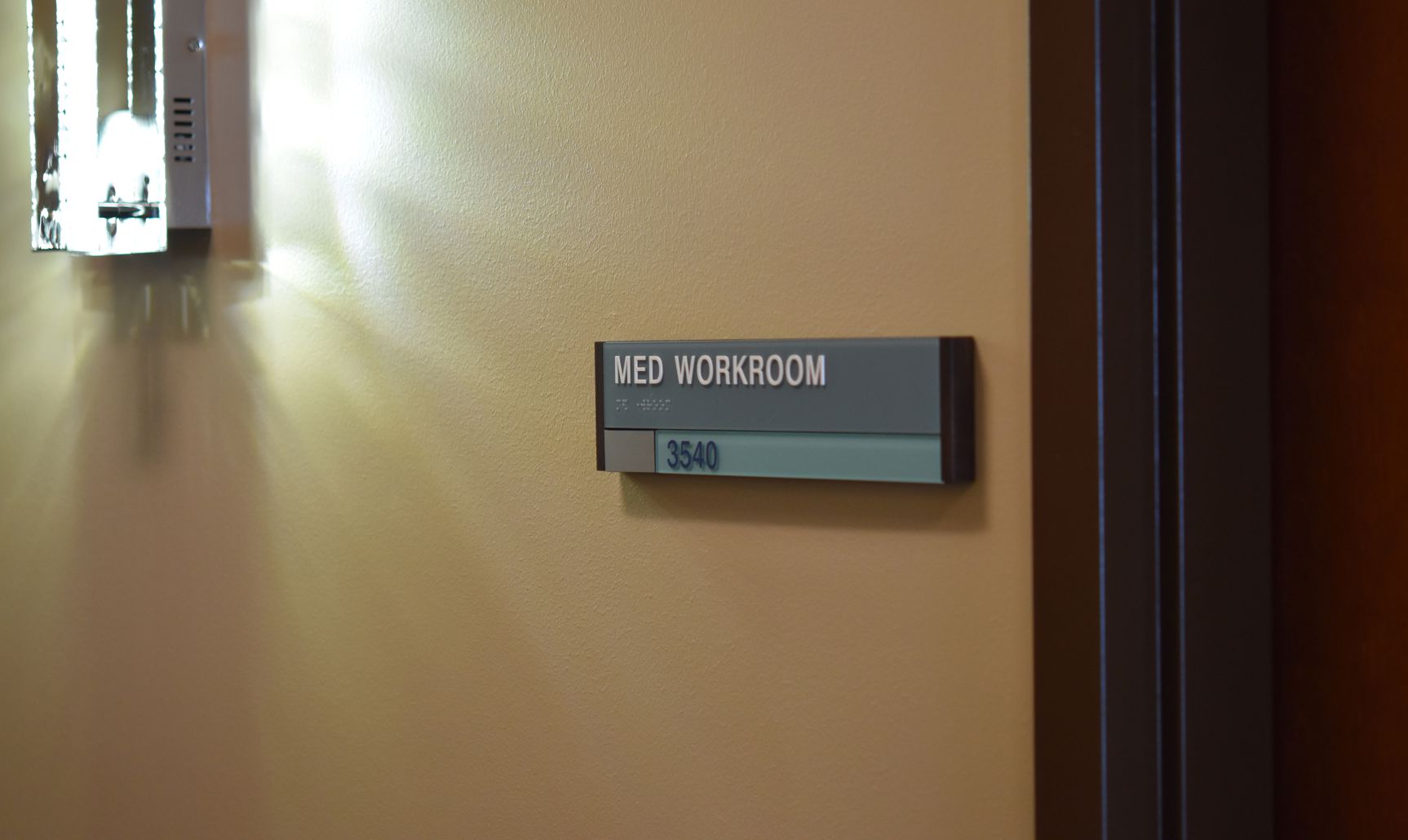 A wall-mounted sign next to a door reads "Med Workroom" with the room number 3540. The sign is gray with white text and braille, and a small portion of the bottom in light blue with the number.
