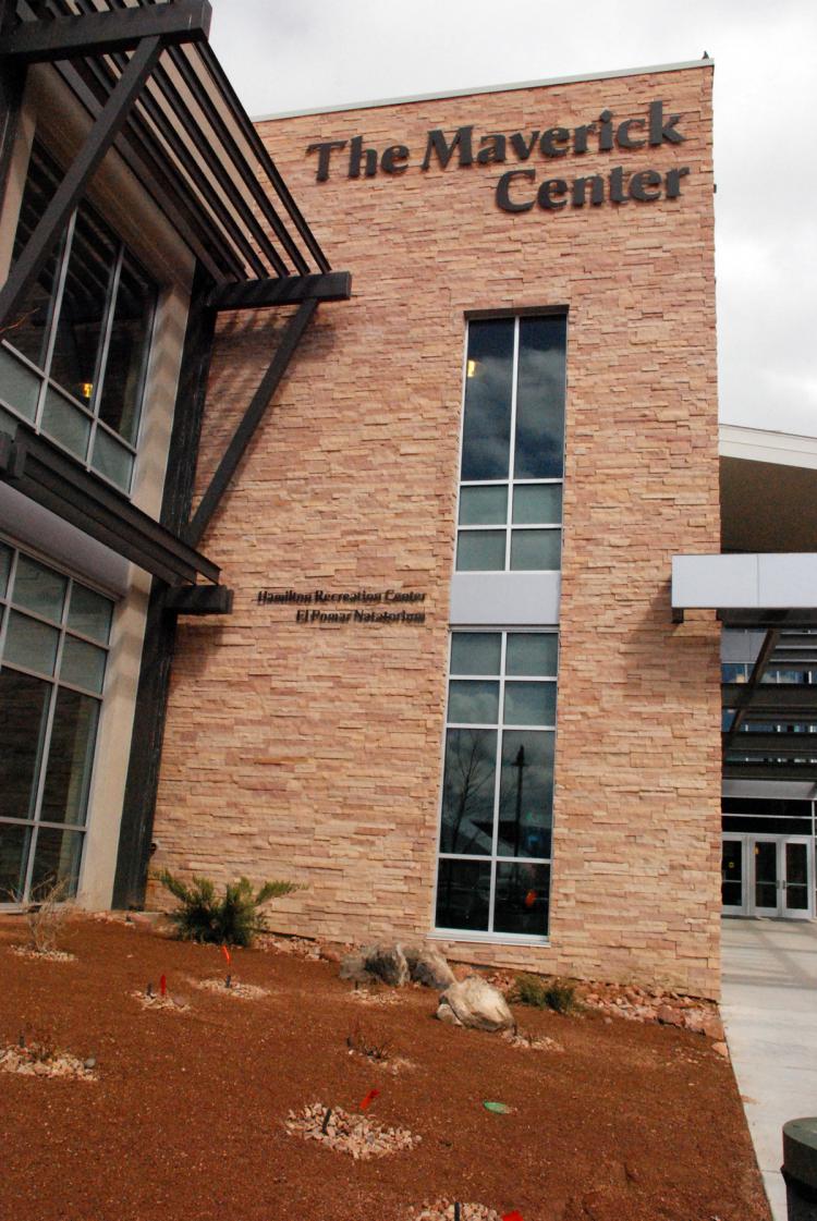 A brick and glass building named "The Maverick Center" with large windows and a modern design is shown. The entrance features an overhang and a paved pathway, surrounded by a lawn and landscaping under a partly cloudy sky.