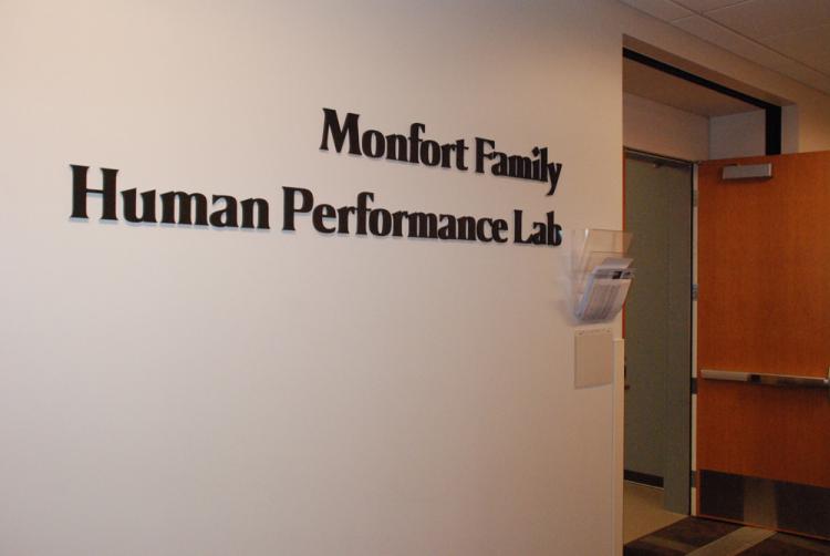 A wall displaying the sign "Monfort Family Human Performance Lab" in black letters. There's a clear wall-mounted brochure holder below the sign. A wooden door stands open to the right, leading into the room. The setting appears to be a professional or academic space.