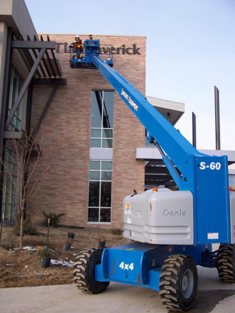 Two workers in safety gear are elevated on a blue Genie S-60 boom lift, performing maintenance or construction on the exterior of a brick building with large windows. Equipment and materials are scattered on the ground below.