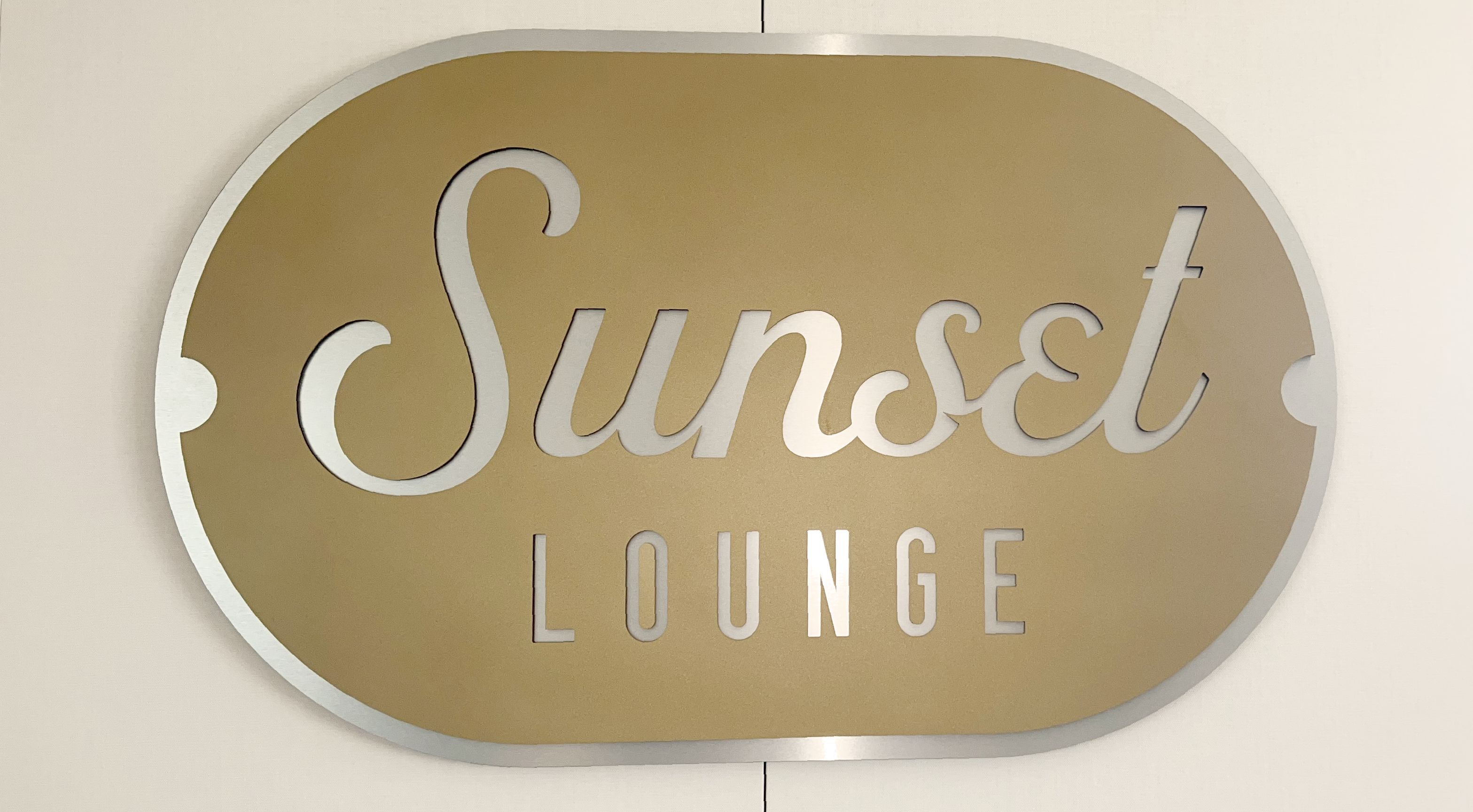 A metallic sign on a wall reads "Sunset Lounge" in elegant script. The background of the sign is a reflective silver color. 
