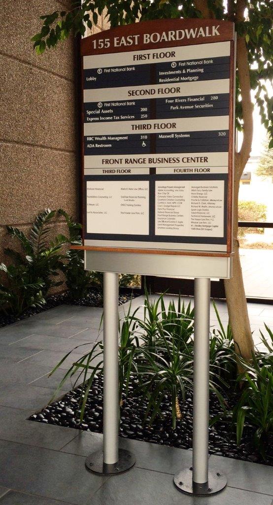 Directory sign at 155 East Boardwalk showing floor listings for various businesses, standing in an indoor setting with surrounding potted plants and a background of windows and railings.