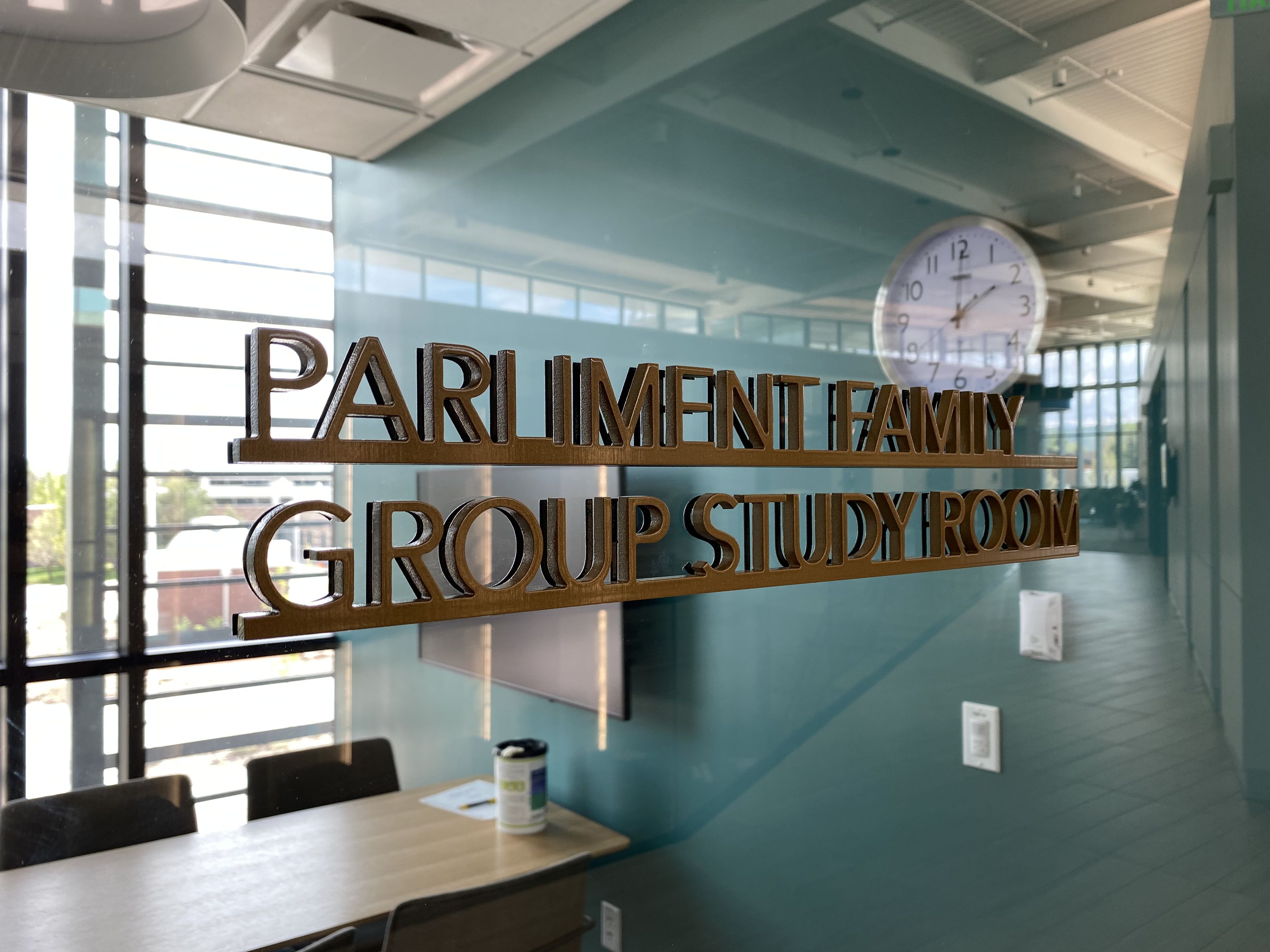 A glass door with a sign that reads "Parliament Family Group Study Room." Inside the room, there is a table with chairs, a reusable water bottle on the table, a clock on the wall, and large windows allowing natural light to enter.