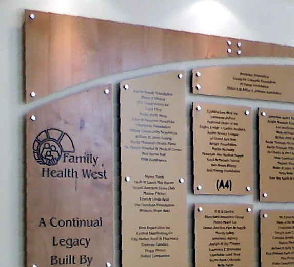 A wooden plaque with metal accents displays multiple engraved names and organizations under the heading "Family Health West." The plaque's logo is on the left, and the text below reads "A Continual Legacy Built By." The plaque is mounted on a light wall.