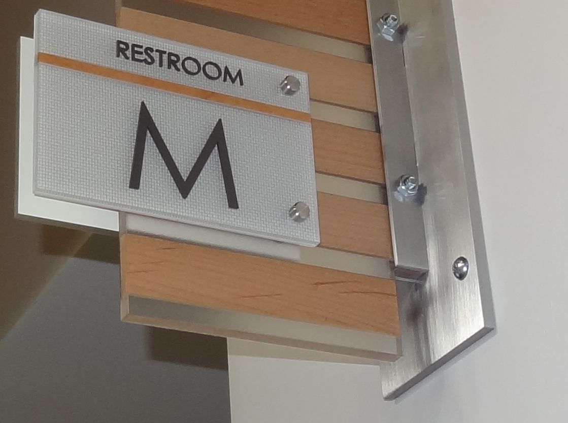 A restroom sign mounted on a wall indicates the men's restroom with a letter "M". The sign is attached to a wooden panel with horizontal slats. The word "RESTROOM" appears above the "M" on the sign.