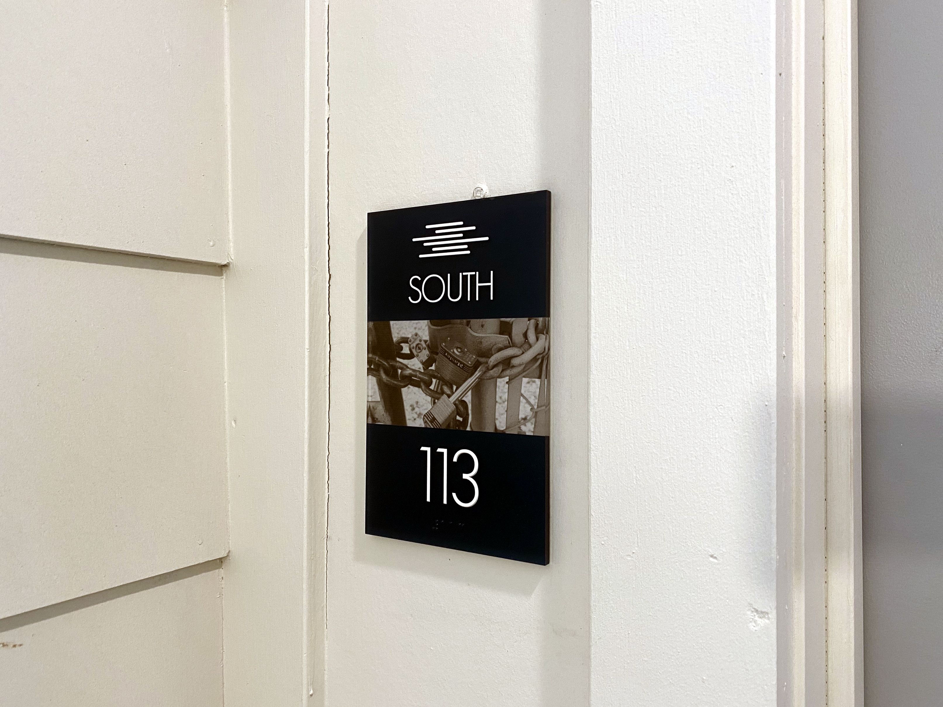 A black and white wall sign displays the text "SOUTH 113" with a stylized horizontal line design. It's mounted beside a beige, paneled door, and to the right is a plain white wall. The background image on the sign shows a black and white photo.