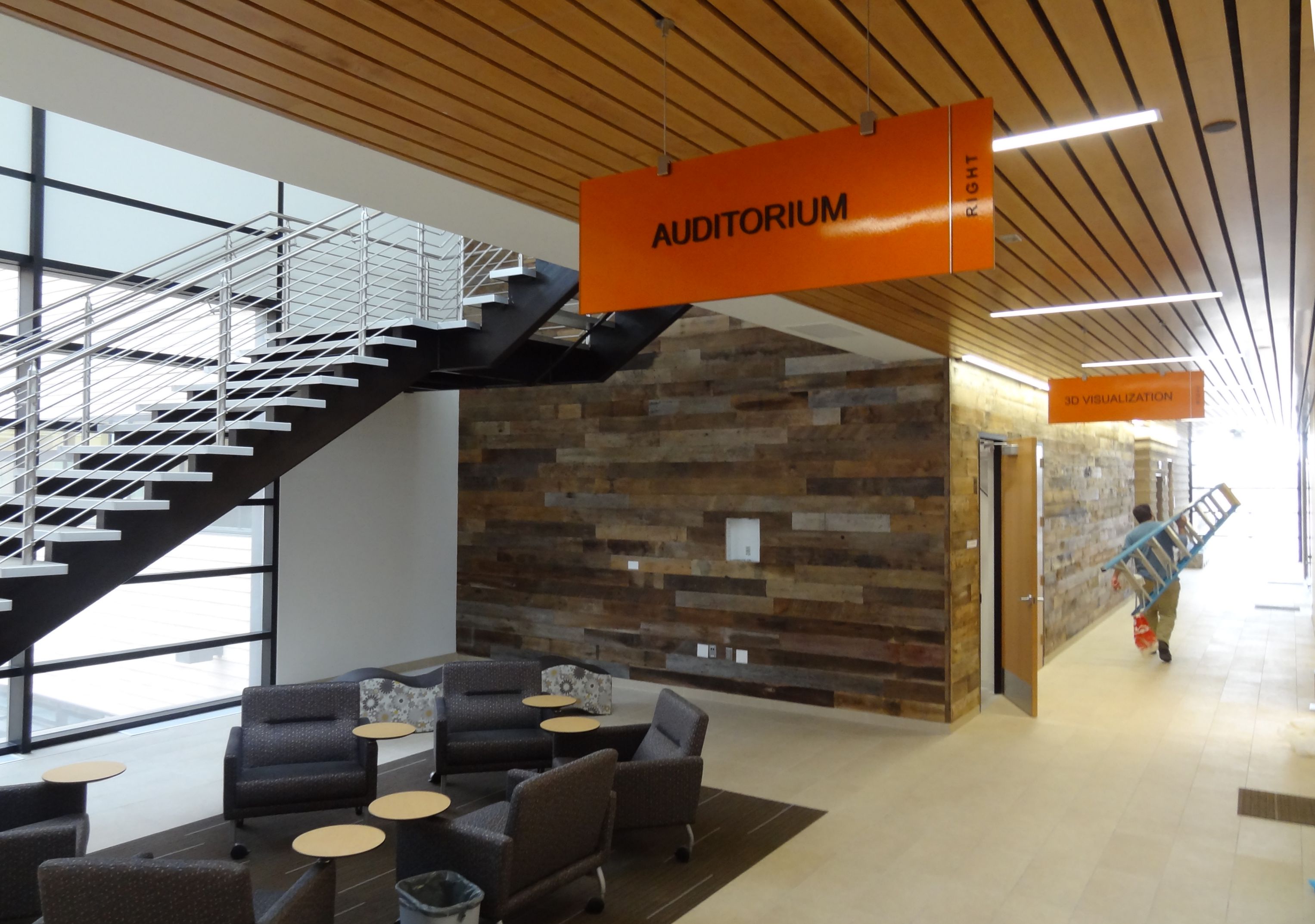 A modern indoor space with a wooden ceiling and walls. There's a staircase with metal rails, a few grey lounge chairs, small tables, and orange signs indicating directions to an auditorium and visual exhibition. A man carrying a ladder is visible in the background.