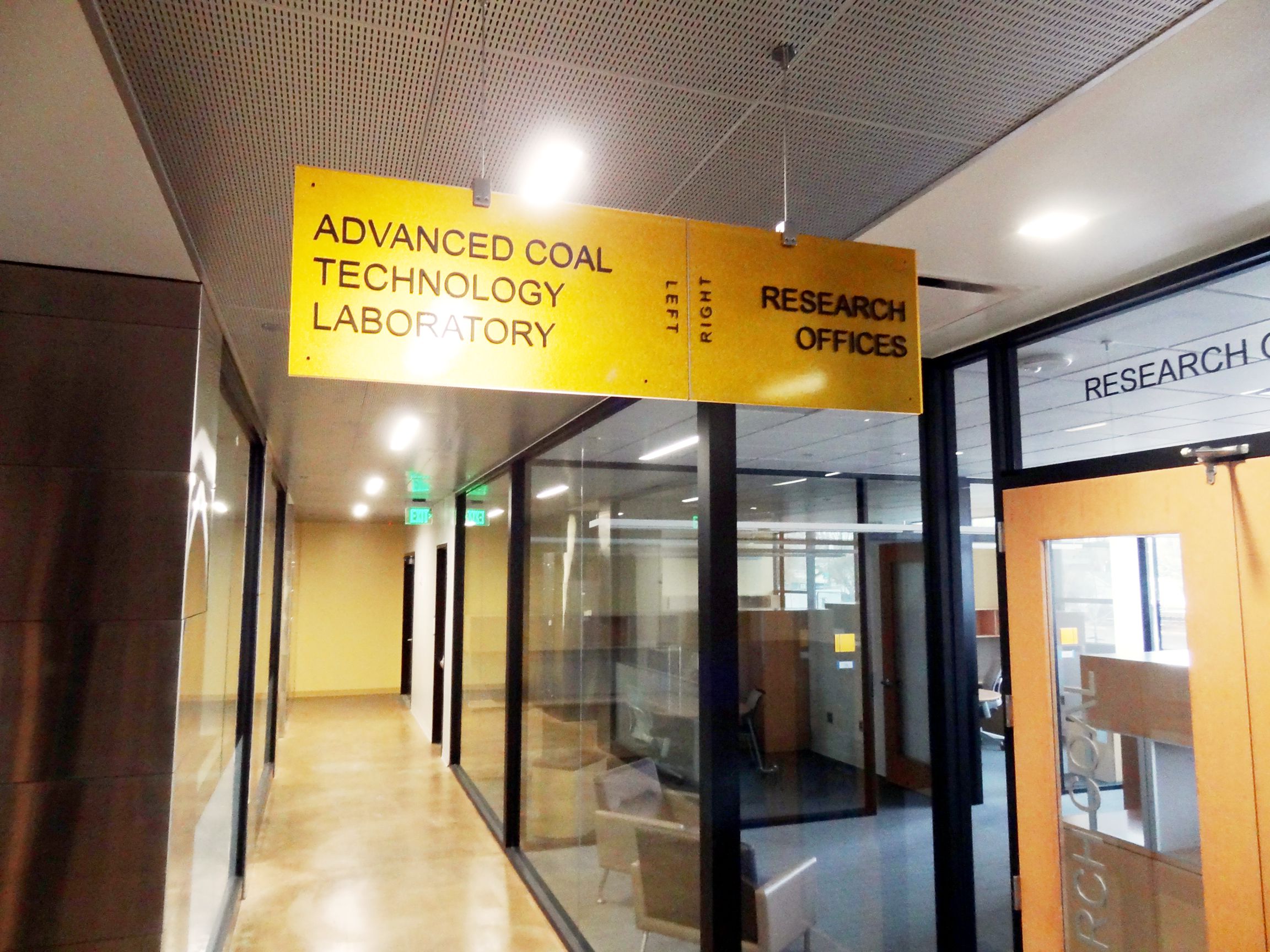A hallway in a building features a yellow sign hanging from the ceiling that reads "Advanced Coal Technology Laboratory" with a right arrow, and "Research Offices" with a left arrow. The hallway leads to glass doors and offices labeled "Research Office.