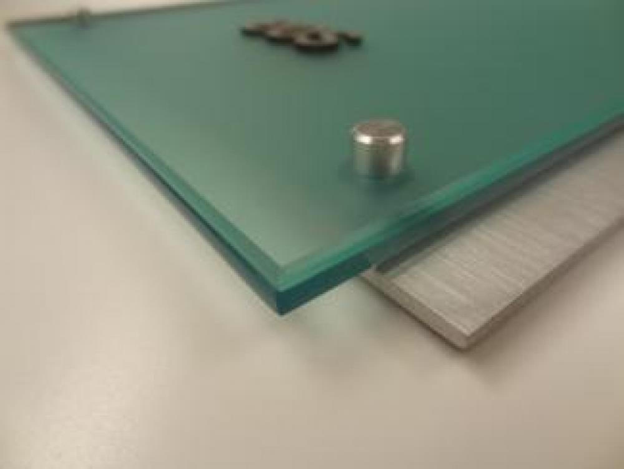 Close-up of a corner of a modern glass sign mounted on a brushed metal backplate. The glass sign is held in place by cylindrical metal standoffs, giving it a floating appearance. The surface of the glass is smooth and has a slight greenish tint.
