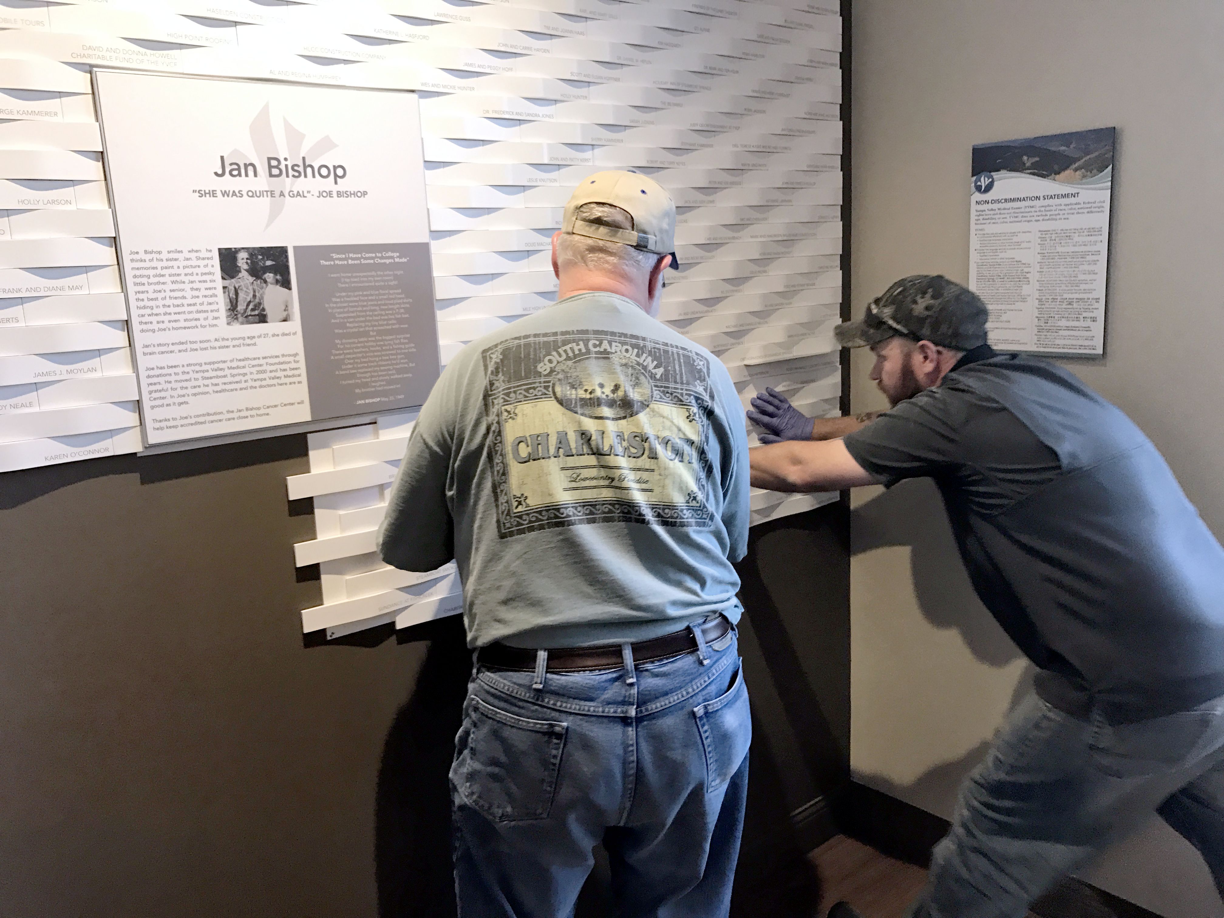 Two men are mounting a wall display featuring information about Jan Bishop. The display includes text and a photograph of Jan Bishop.