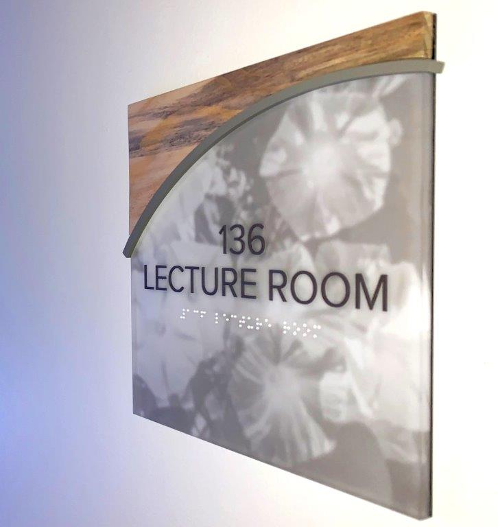 A room sign on a wall reads "136 Lecture Room" in large text. Below, smaller Braille text is visible. The sign has a grey, semi-transparent background with floral patterns and a wooden accent at the top.
