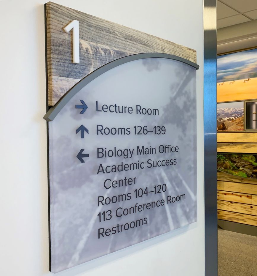 A wall sign on the first floor directs to a lecture room, rooms 126-139, the Biology Main Office, Academic Success Center, rooms 104-120, and a conference room (113). The sign uses a mix of blue and black text with arrows beside each listing.