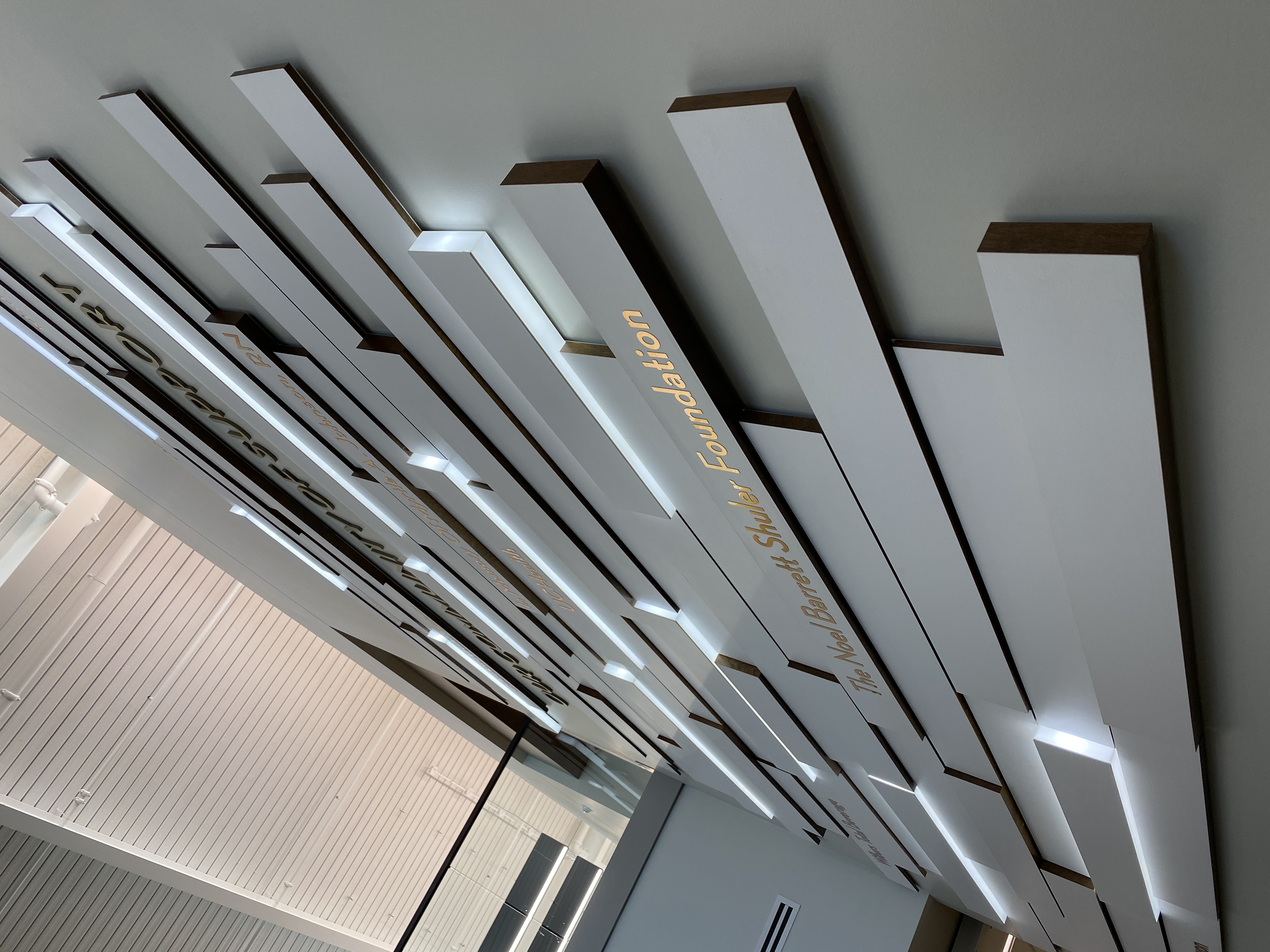 A wall display titled "Our Community of Support" with illuminated horizontal plaques in a modern building. Names of donors are visible on each plaque. There's a glass balcony railing above, and the area is well-lit with a neutral color scheme.