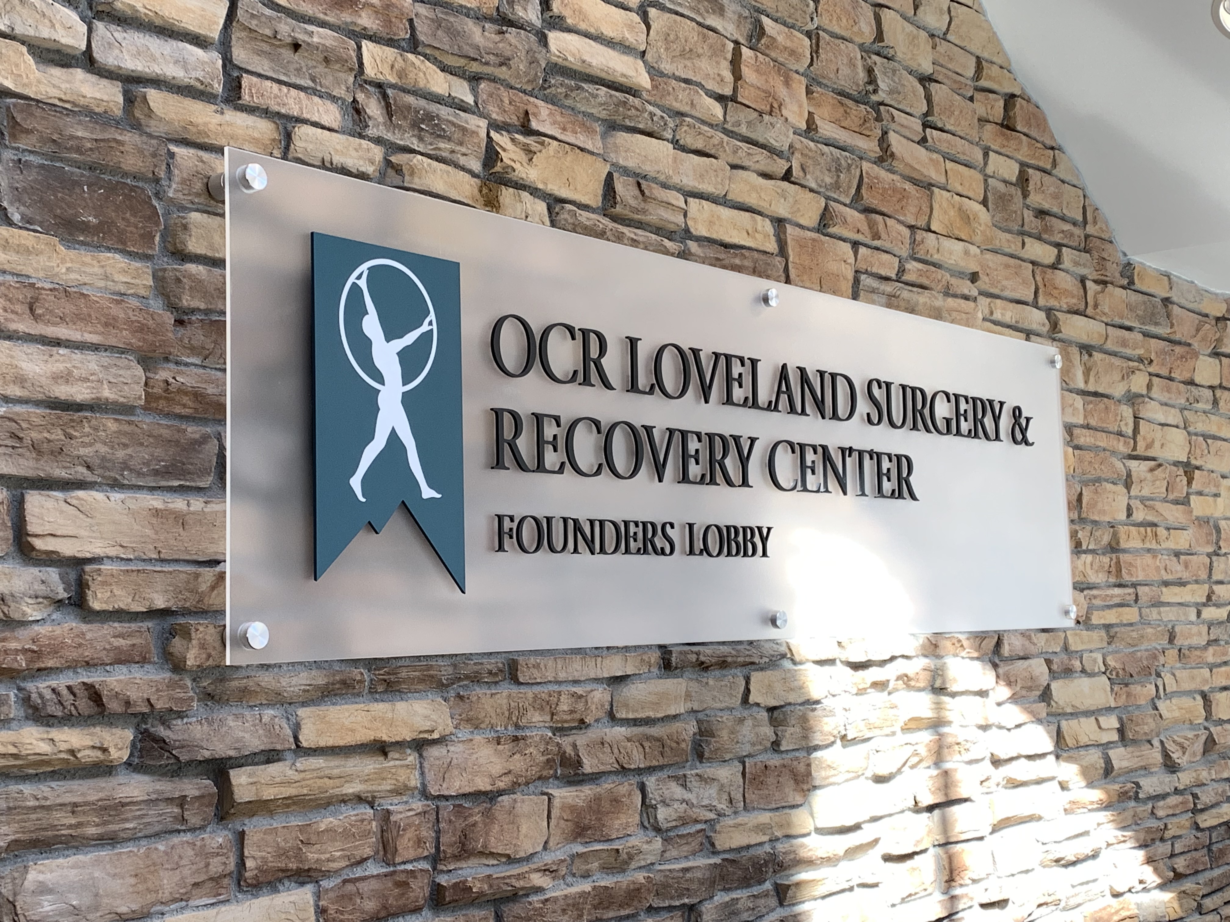 A sign on a brick wall reads "OCR Loveland Surgery & Recovery Center, Founders Lobby." It features a blue emblem with a white silhouette of a person.