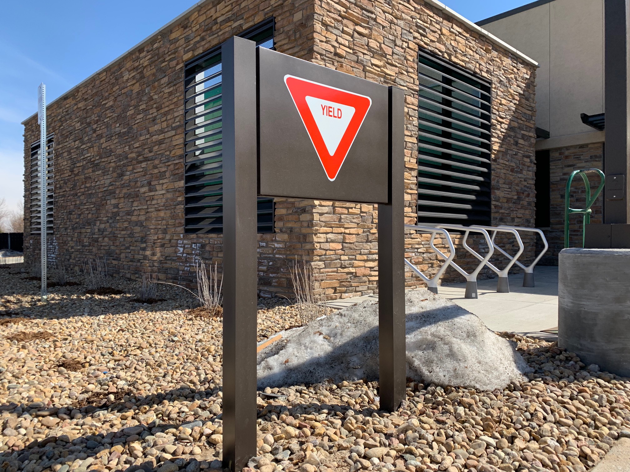 A yield sign mounted on a metal frame stands in front of a modern building with stone exterior walls and large, horizontal window slats. The ground is covered with small pebbles and some patches of melted snow. A bike rack is visible near the building entrance.