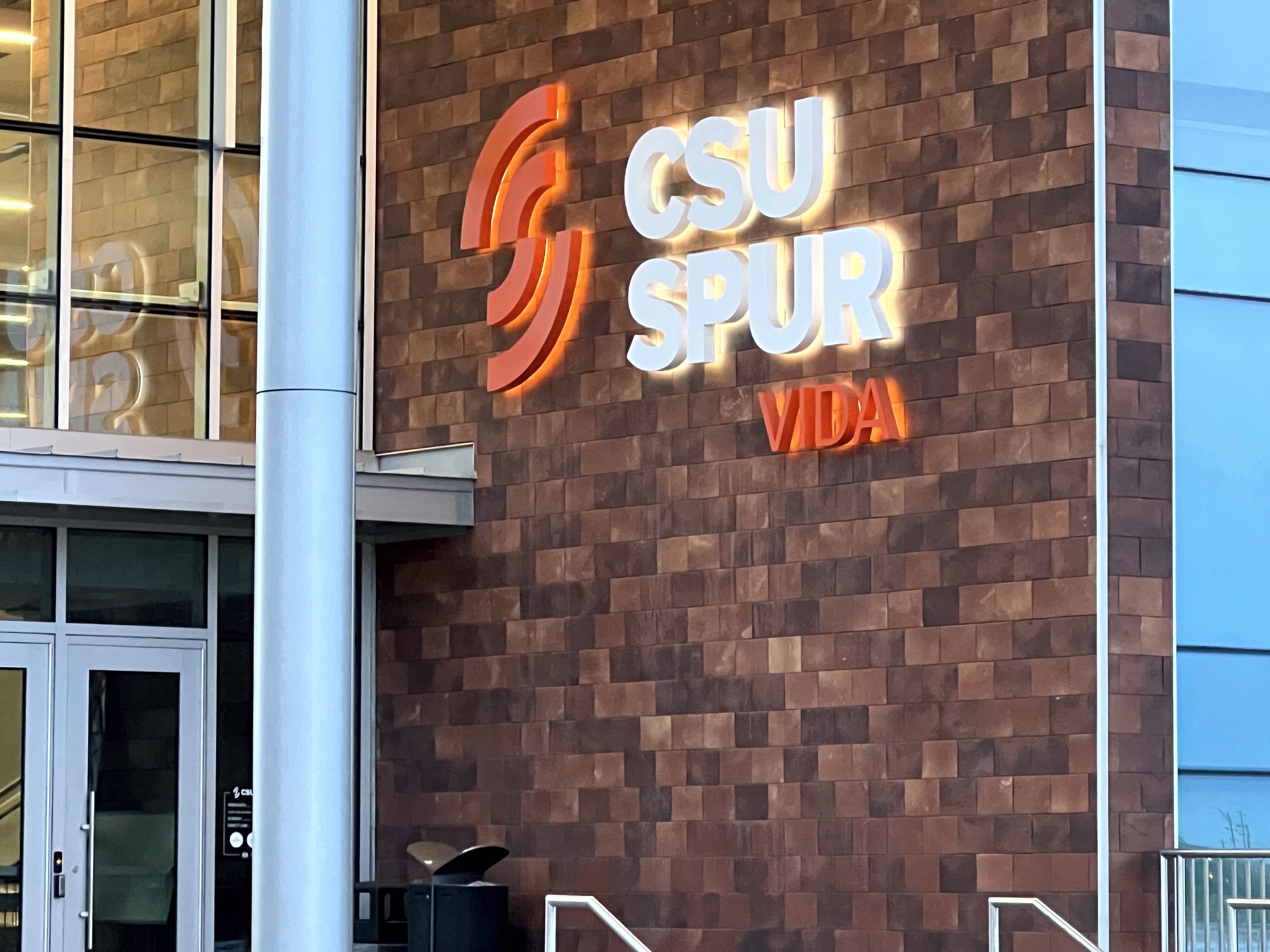 The image shows a wall sign with the text "CSU SPUR VIDA" illuminated. The letters "CSU" and "SPUR" are in white, while the word "VIDA" is in red. The wall is made of brown rectangular tiles, and the letters are outlined with a glowing effect.