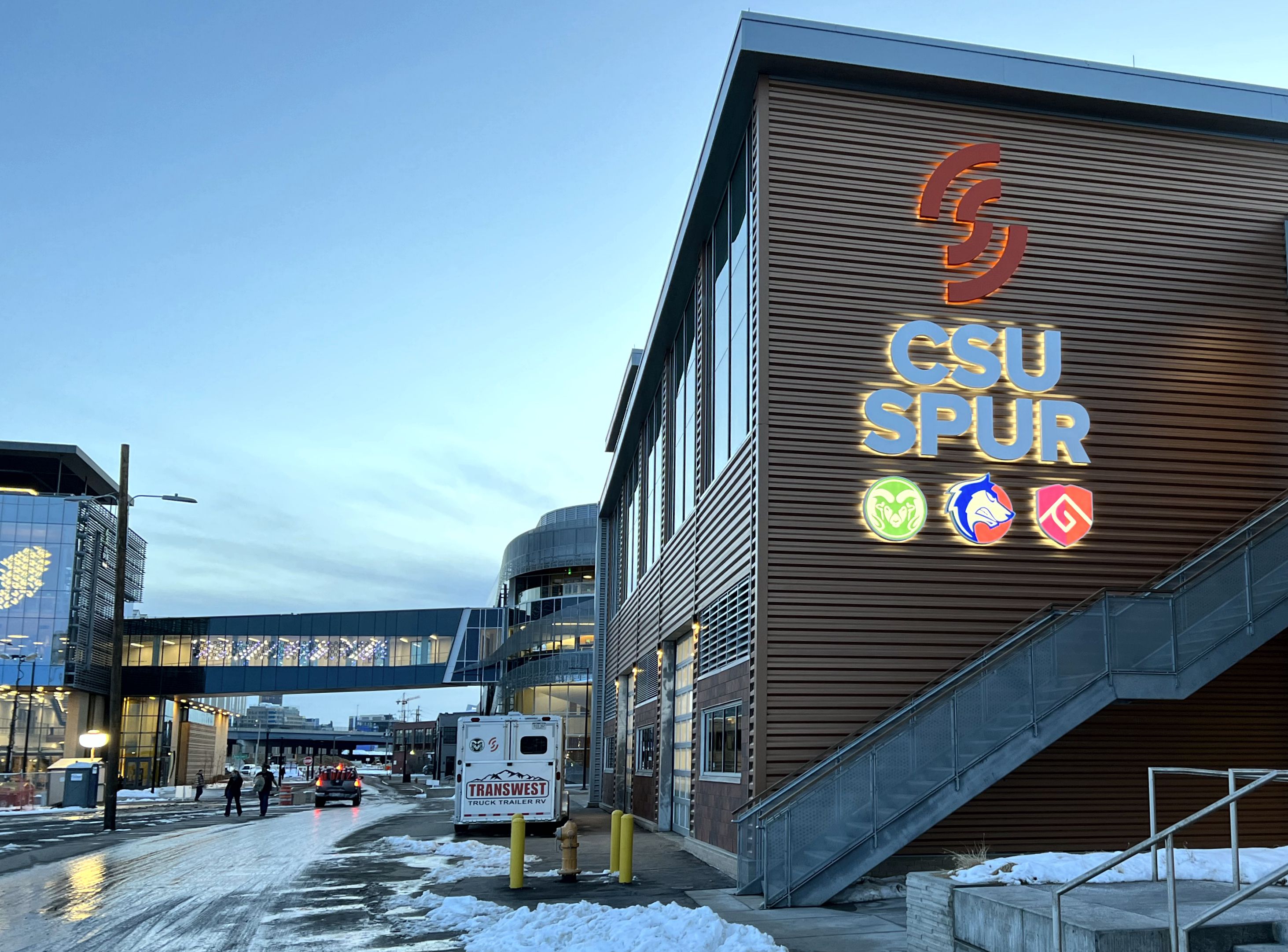 A modern building with large "CSU SPUR" sign and three illuminated logos on its facade. Snow covers the ground and an overpass bridge is visible. 