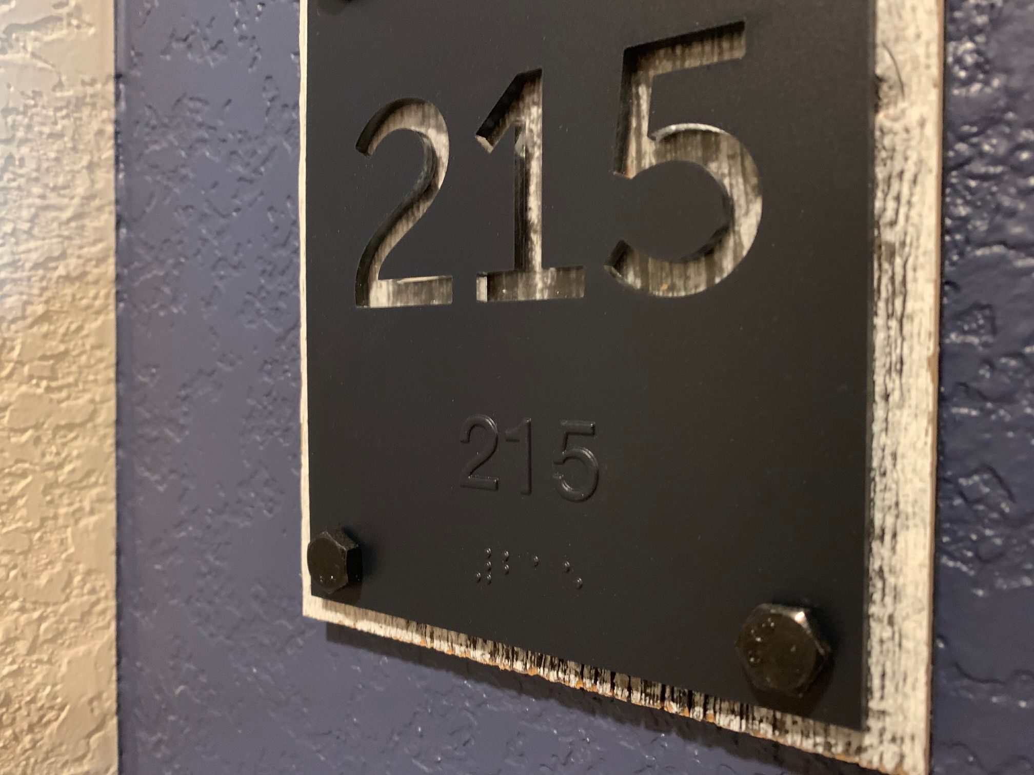 A close-up of a room number sign on a textured blue and beige wall. The sign has the number "215" cut out, with the same number printed below in Braille. The sign is secured with bolts at the corners.