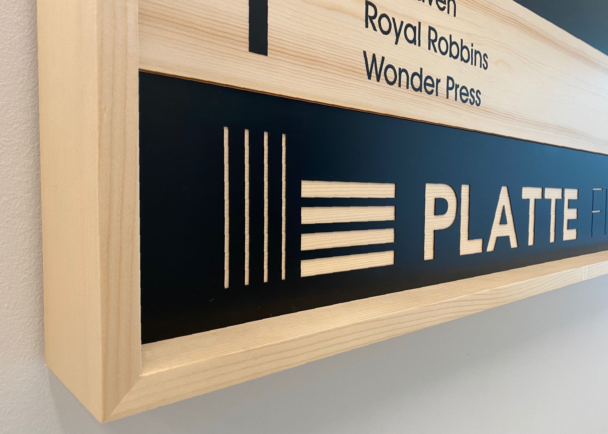 Close-up of a wooden directory sign with engraved horizontal lines and text. The sign features the word "PLATTE" in white letters on a black background, bordered by light wood. Part of the directory listing is visible above.