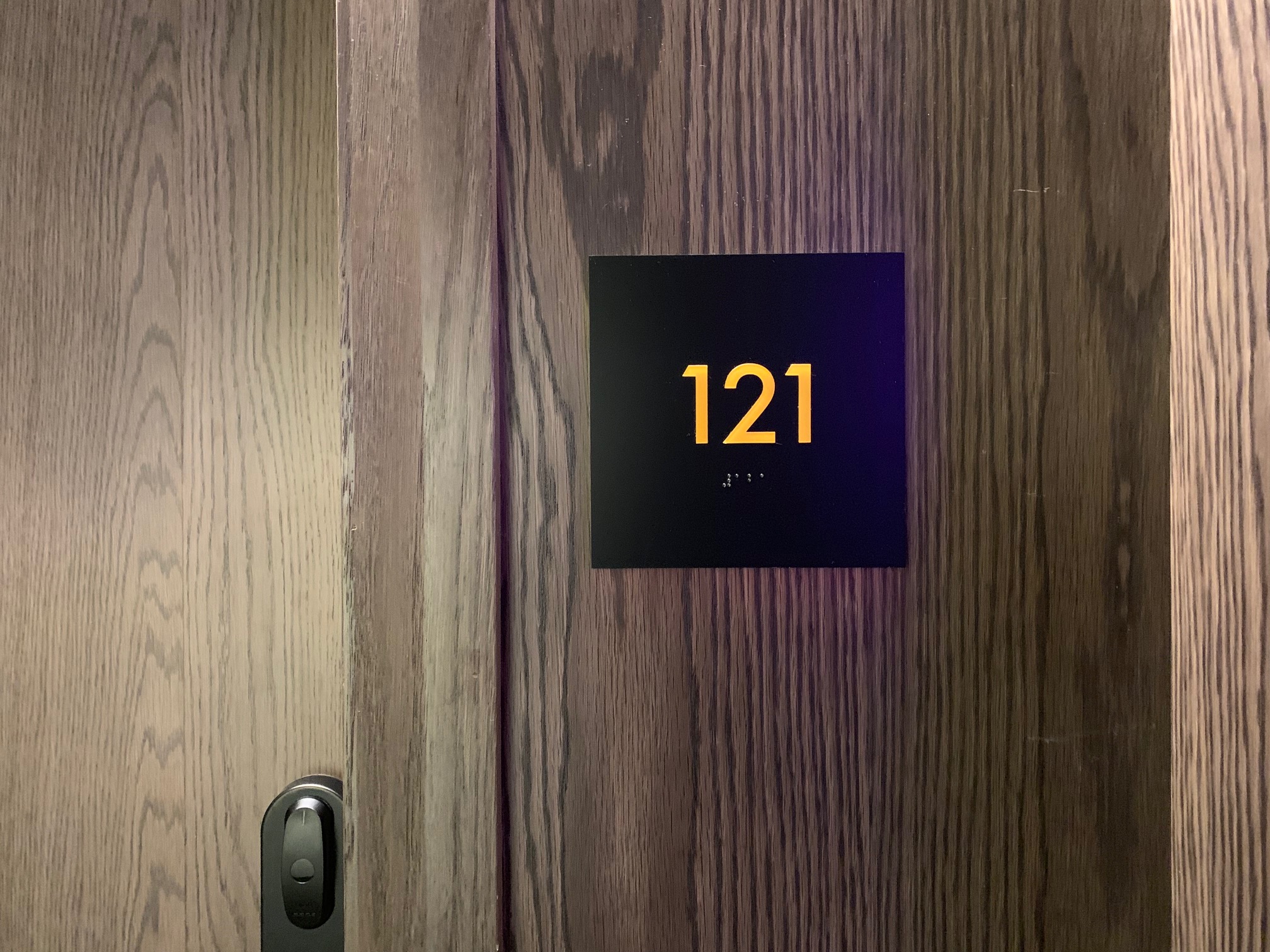 A close-up of a dark wooden door with a black rectangular plaque displaying the number "121" in yellow. The sign also includes Braille beneath the numbers.