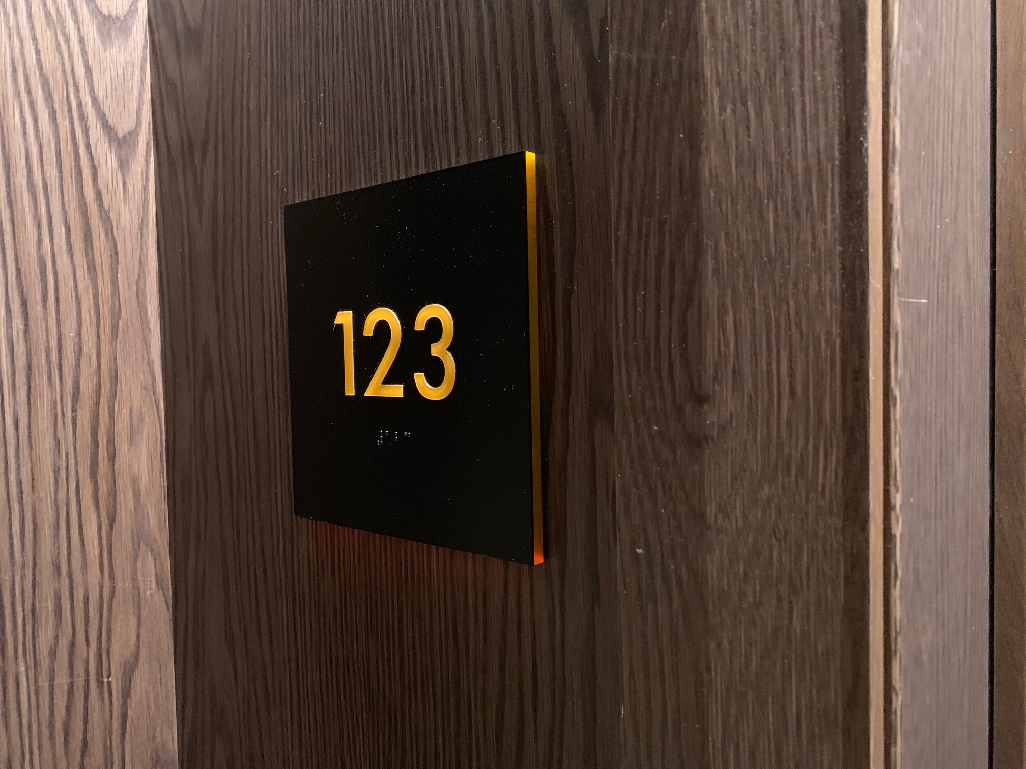 A close-up of a dark wooden door with a black rectangular plaque displaying the number "123" in yellow. The sign also includes Braille beneath the numbers.