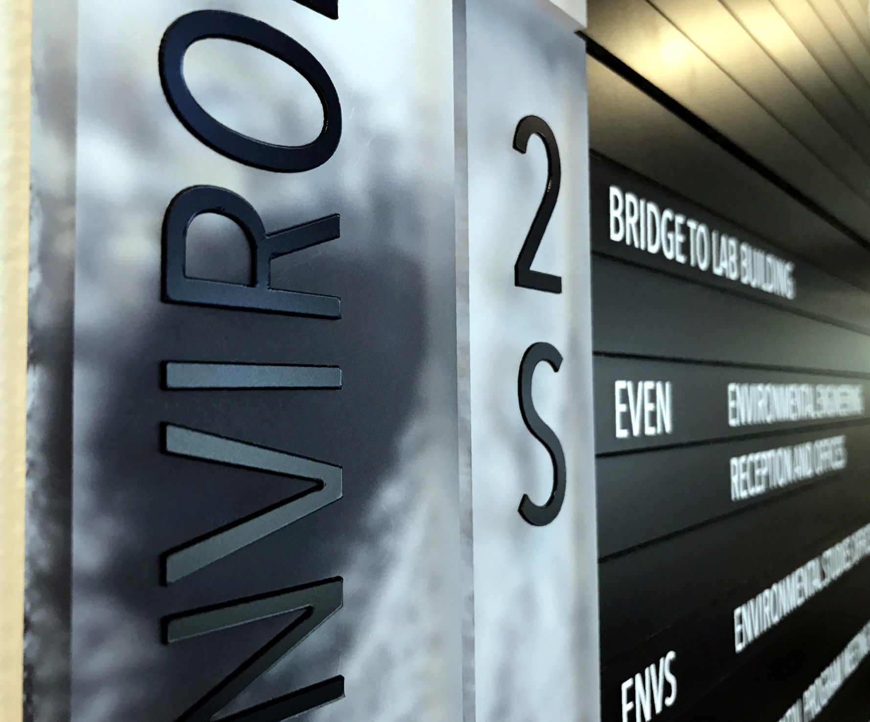 Close-up of a building directory sign with the word "ENVIRONMENT" vertically displayed on a frosted glass panel on the left. The right side shows directions to various locations, including "Bridge to Lab Building" and other blurred listings.