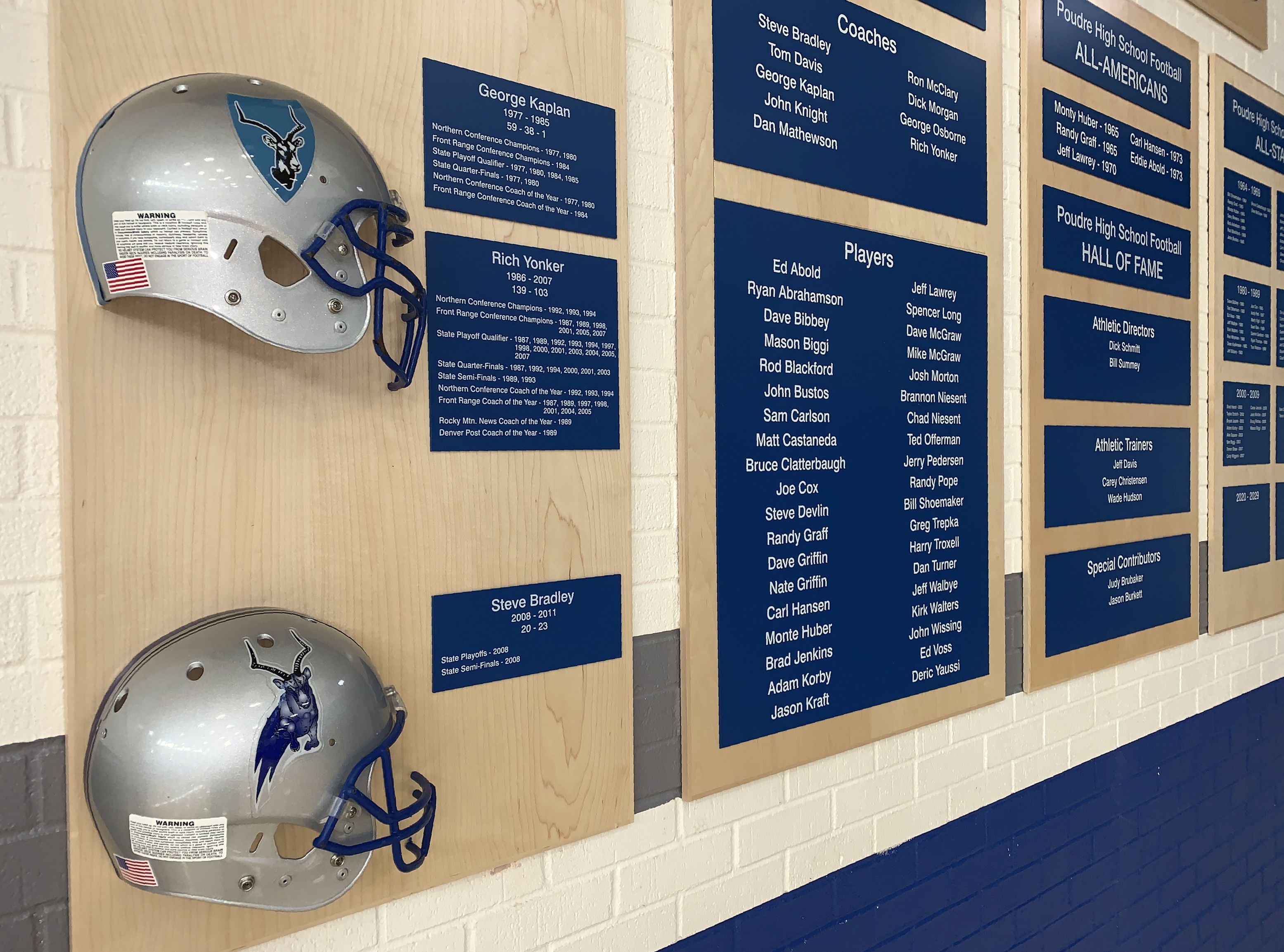 A display on a white tiled wall features two silver football helmets with blue accents mounted on wooden plaques. Adjacent to the helmets are blue information boards listing coaches, players, and hall of fame inductees associated with the football program.