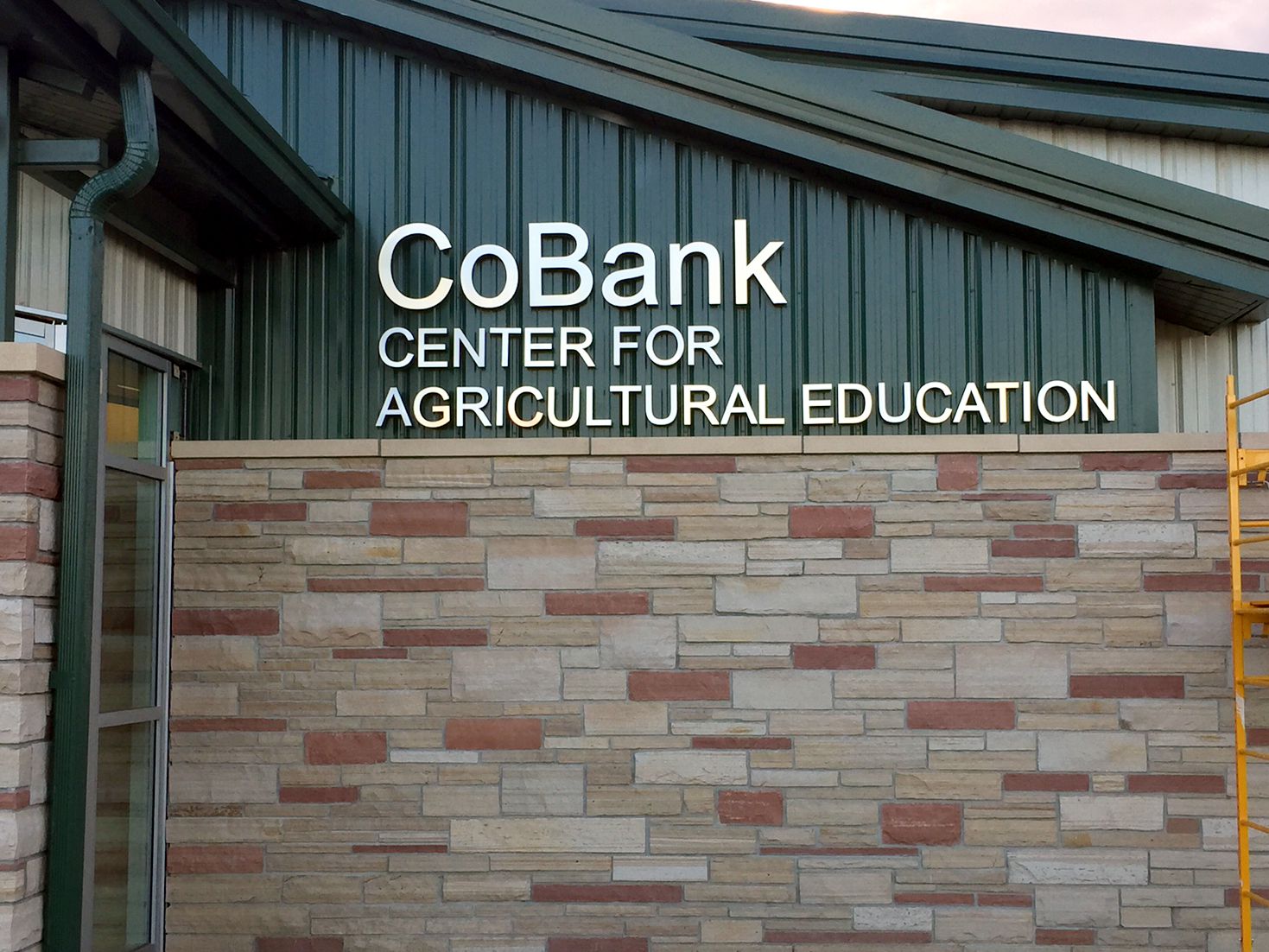 A building with a stone facade and green metal roof displays a sign that reads "CoBank Center for Agricultural Education." A door is partially visible on the left, and to the right, there is a ladder leaning against the wall.