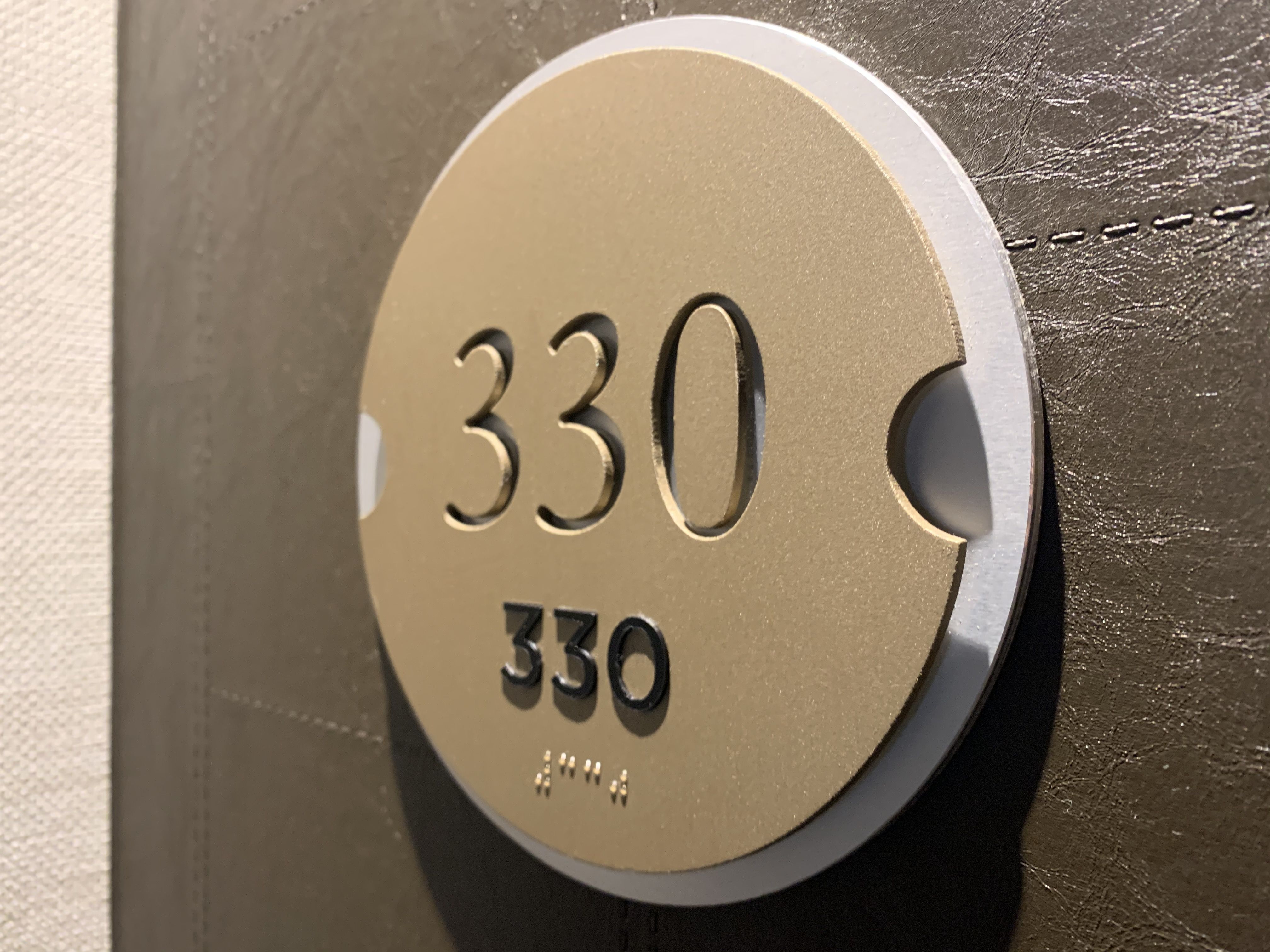 A round beige sign with the number "330" prominently displayed in black numerals is mounted on a gray-tiled wall. The number "330" appears again in smaller print below, with Braille dots below the smaller print.