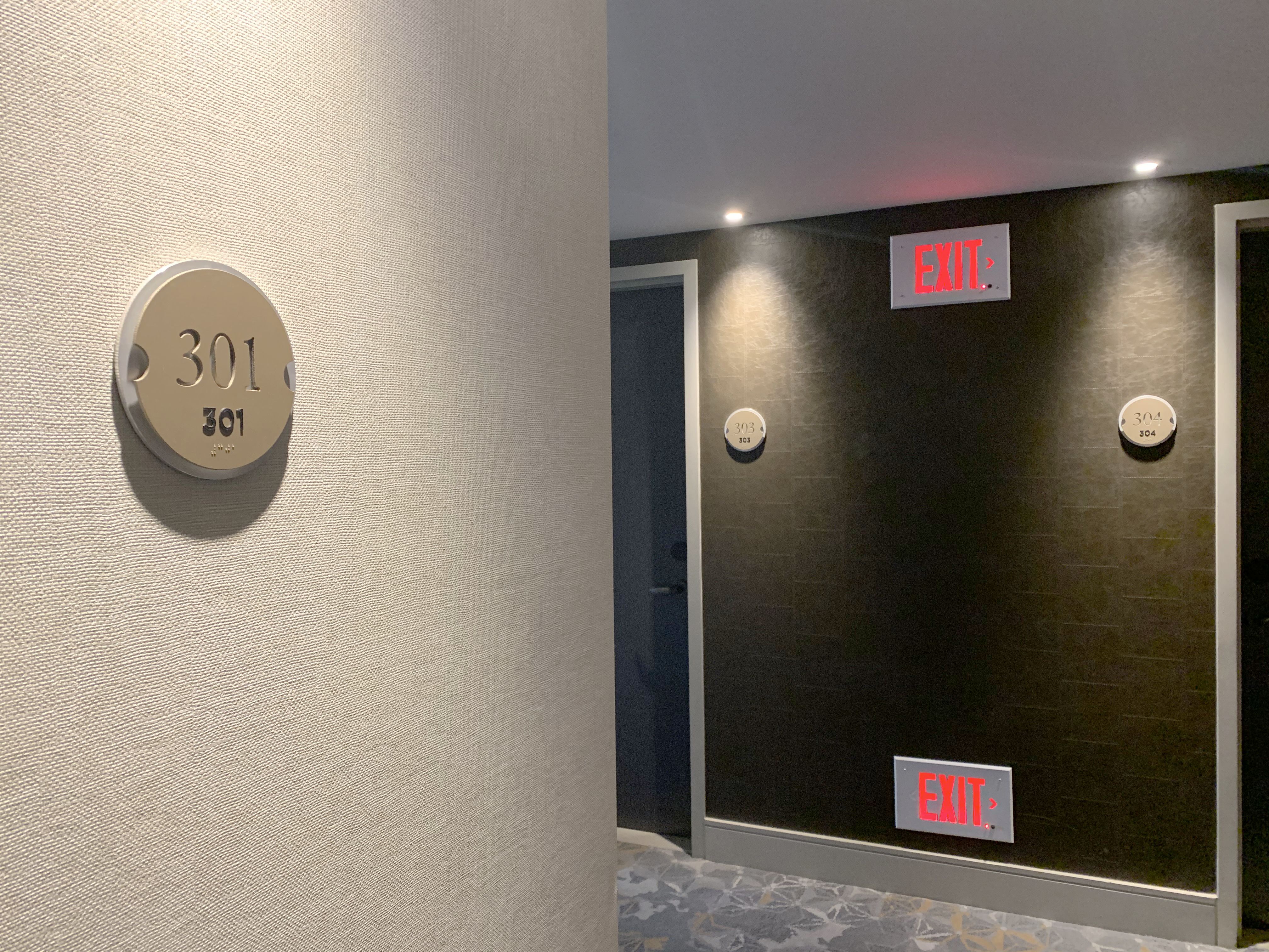 A round beige sign with the number "301" prominently displayed in black numerals is mounted on a gray-tiled wall. The number "301" appears again in smaller print below, with Braille dots below the smaller print.  A hallway with multiple doors and a glowing Exit sign shows in the background.