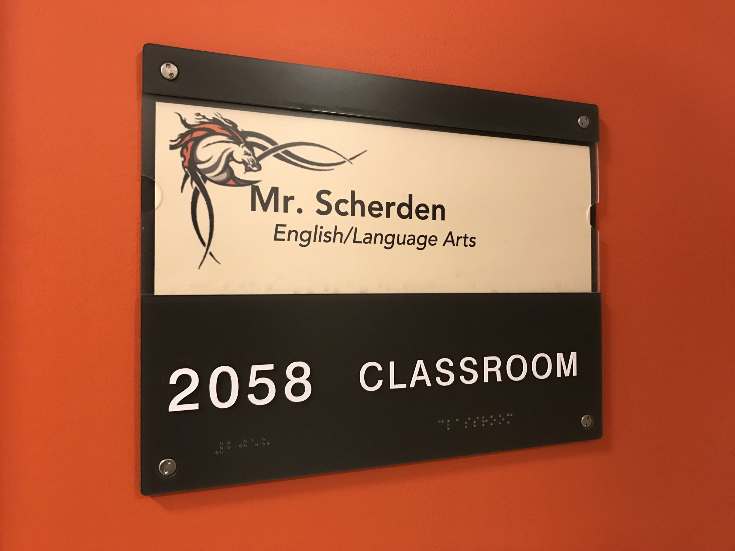 A classroom door sign reads, "Mr. Scherden English/Language Arts" with a stylized stallion image above the name. Below, it indicates the room number "2058" and the word "CLASSROOM." The sign is mounted on an orange wall and includes Braille.