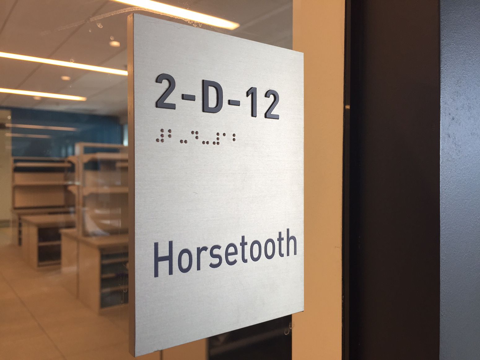 A metallic door sign reads "2-D-12 Horsetooth" in both text and braille. The background shows an office setting with shelves and a glass wall.