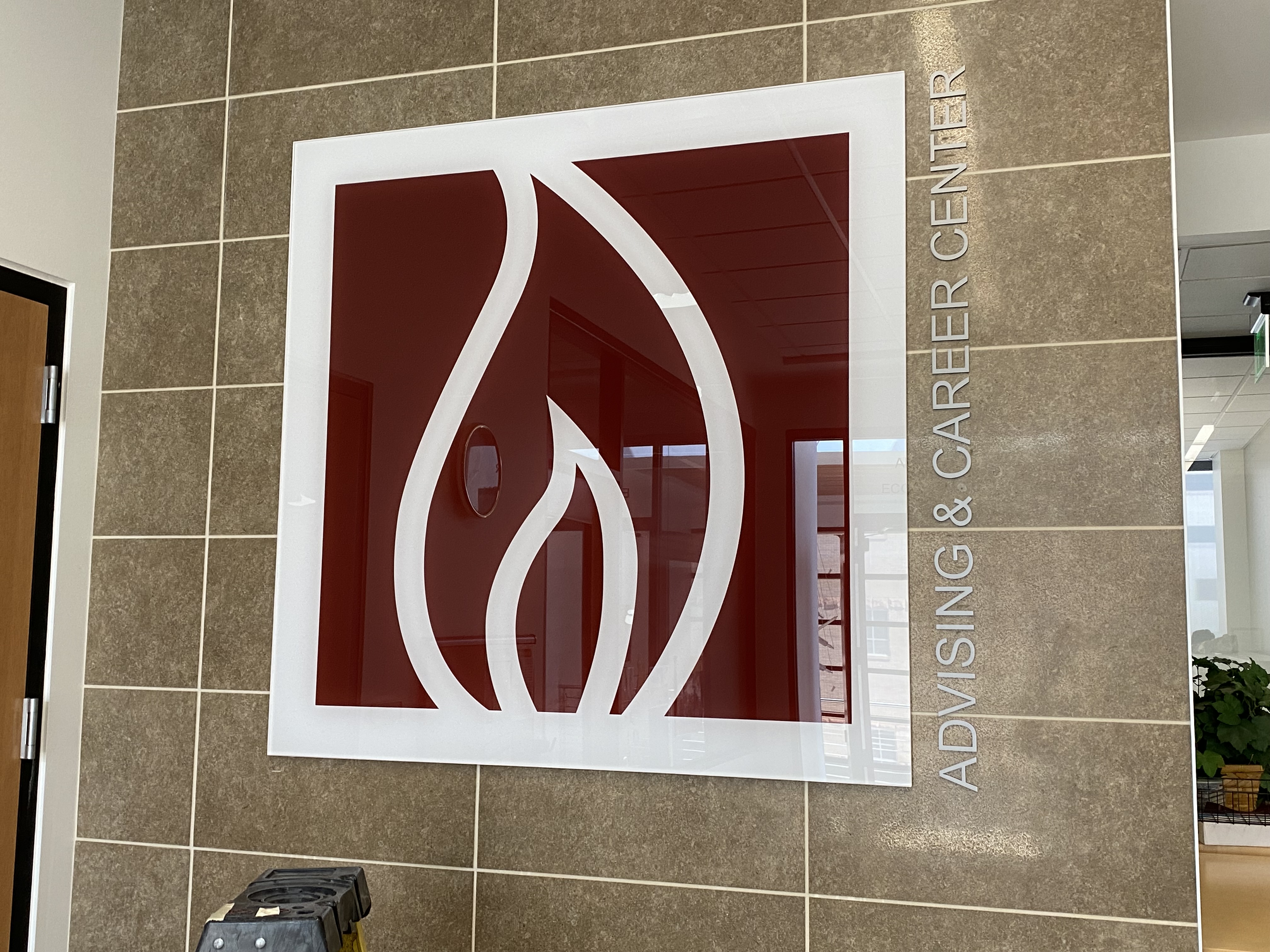 A wall displaying a large, square sign with a stylized flame logo in white against a red background. Next to the sign, the words “Advising & Career Center” are vertically aligned in white text. The surrounding wall is covered in beige tiles.
