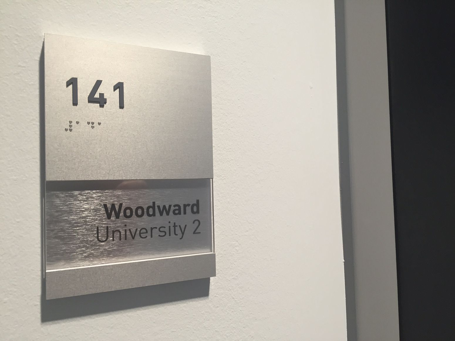 A silver sign on a light-colored wall displays the number "141" with Braille above the text "Woodward University 2" on a gray background. The sign is mounted next to a dark doorway.