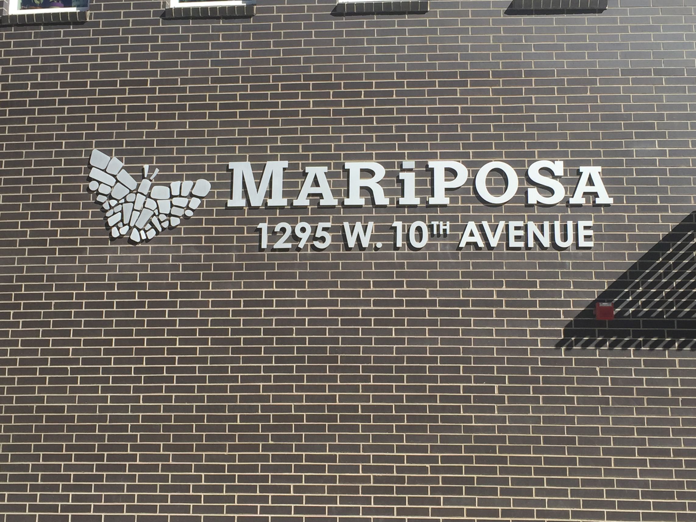 A brick wall with the text "MARIPOSA 1295 W. 10TH AVENUE" in white modern capital letters. To the left of the text is a white butterfly graphic. Shadows and sunlight create a pattern on the wall.