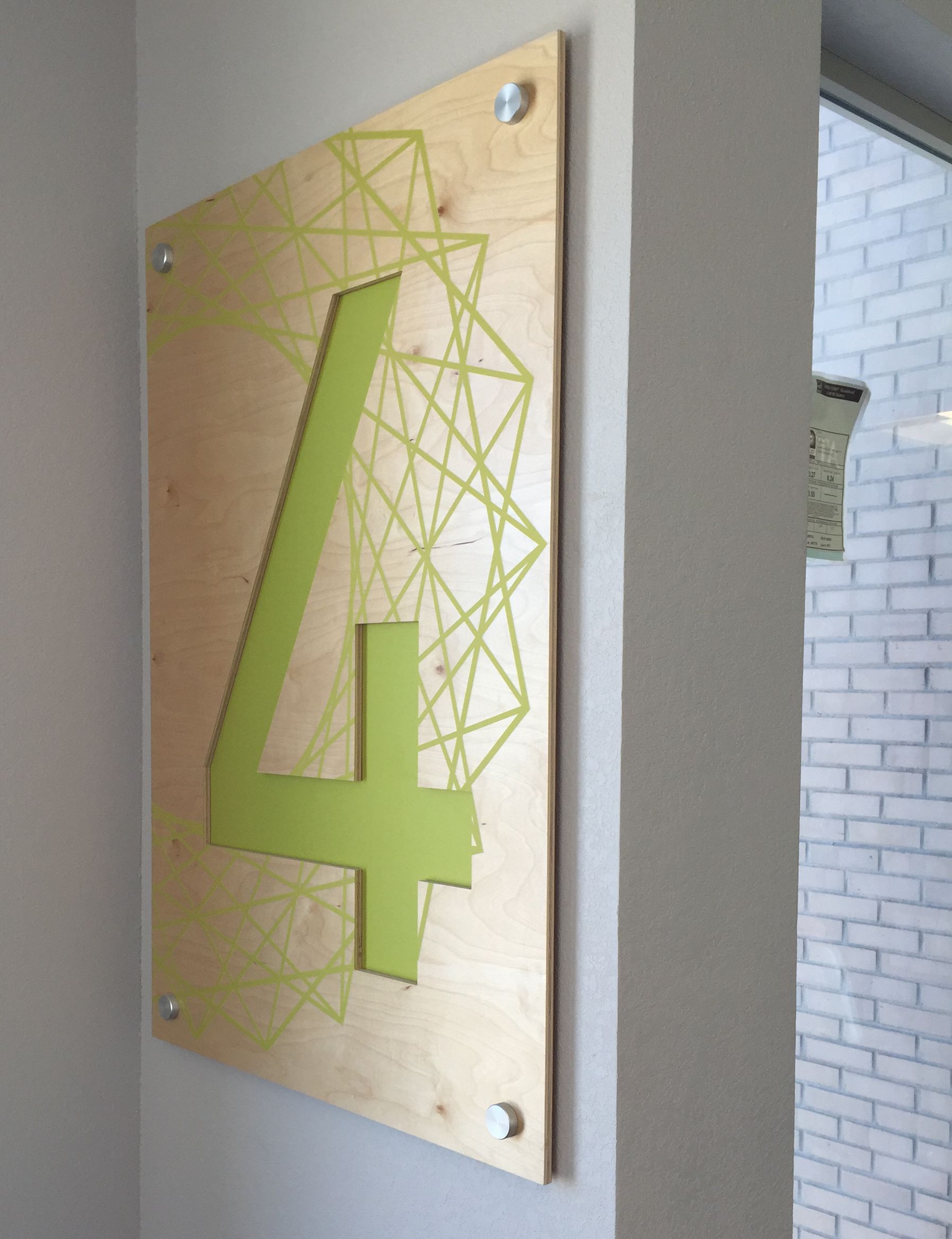 This image shows a wooden wall panel mounted on a wall, marked with a large green numeral "4" and surrounded by a geometric pattern. The panel is affixed with four metallic bolts at each corner. A window, partially visible to the right, reveals a brick wall outside.