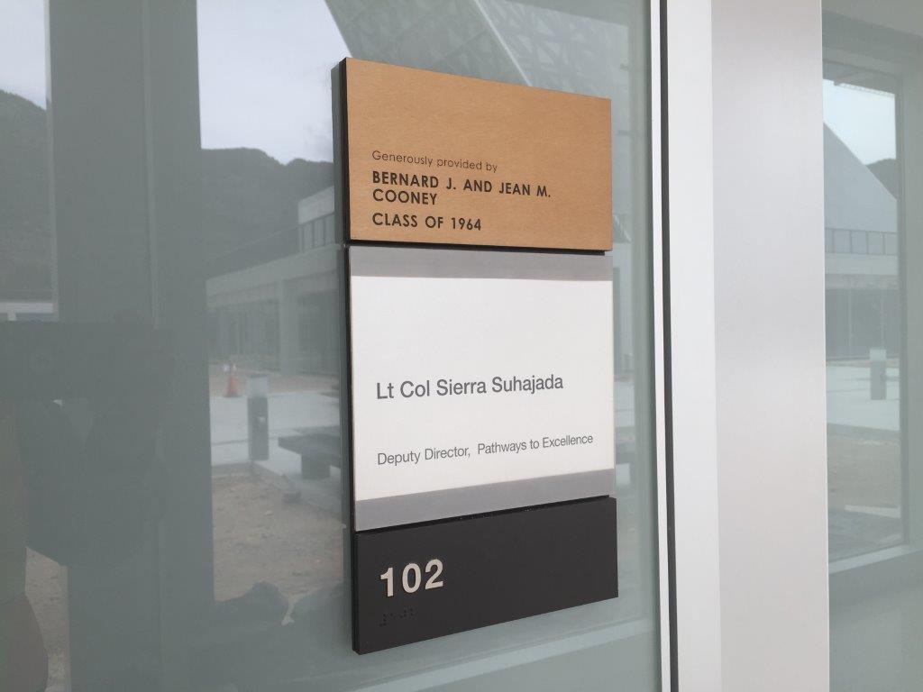 A door sign for an office. The top part reads "Generously provided by Bernard J. and Jean M. Cooney, Class of 1964". Below it, another part reads "Lt Col Sierra Suhajada, Deputy Director, Pathways to Excellence". The bottom part shows the room number "102".