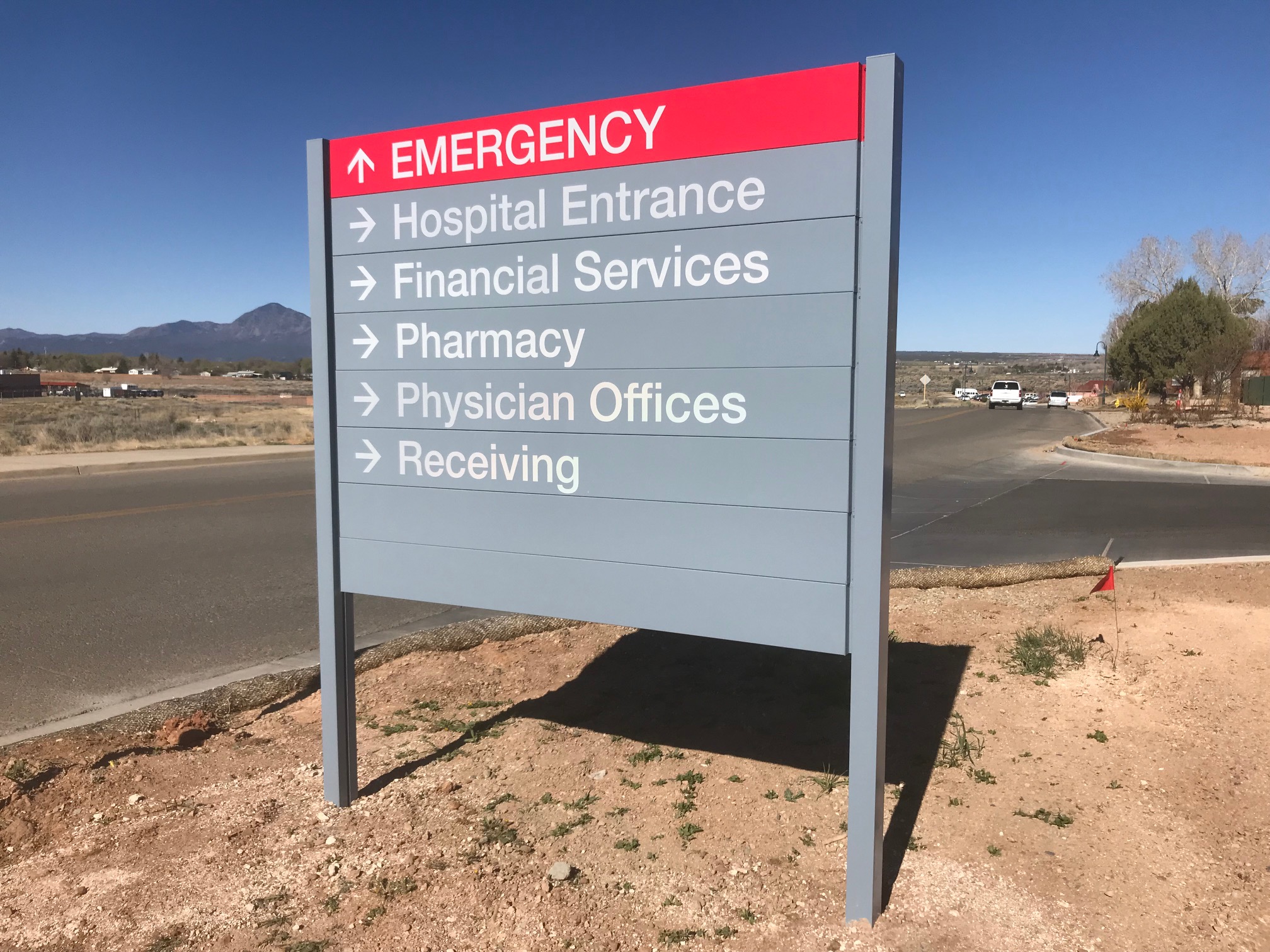A directional sign in a rural area provides guidance for a hospital, including Emergency, Hospital Entrance, Financial Services, Pharmacy, Physician Offices, and Receiving. The sign has a red emergency indicator at the top, and a road is visible in the background.