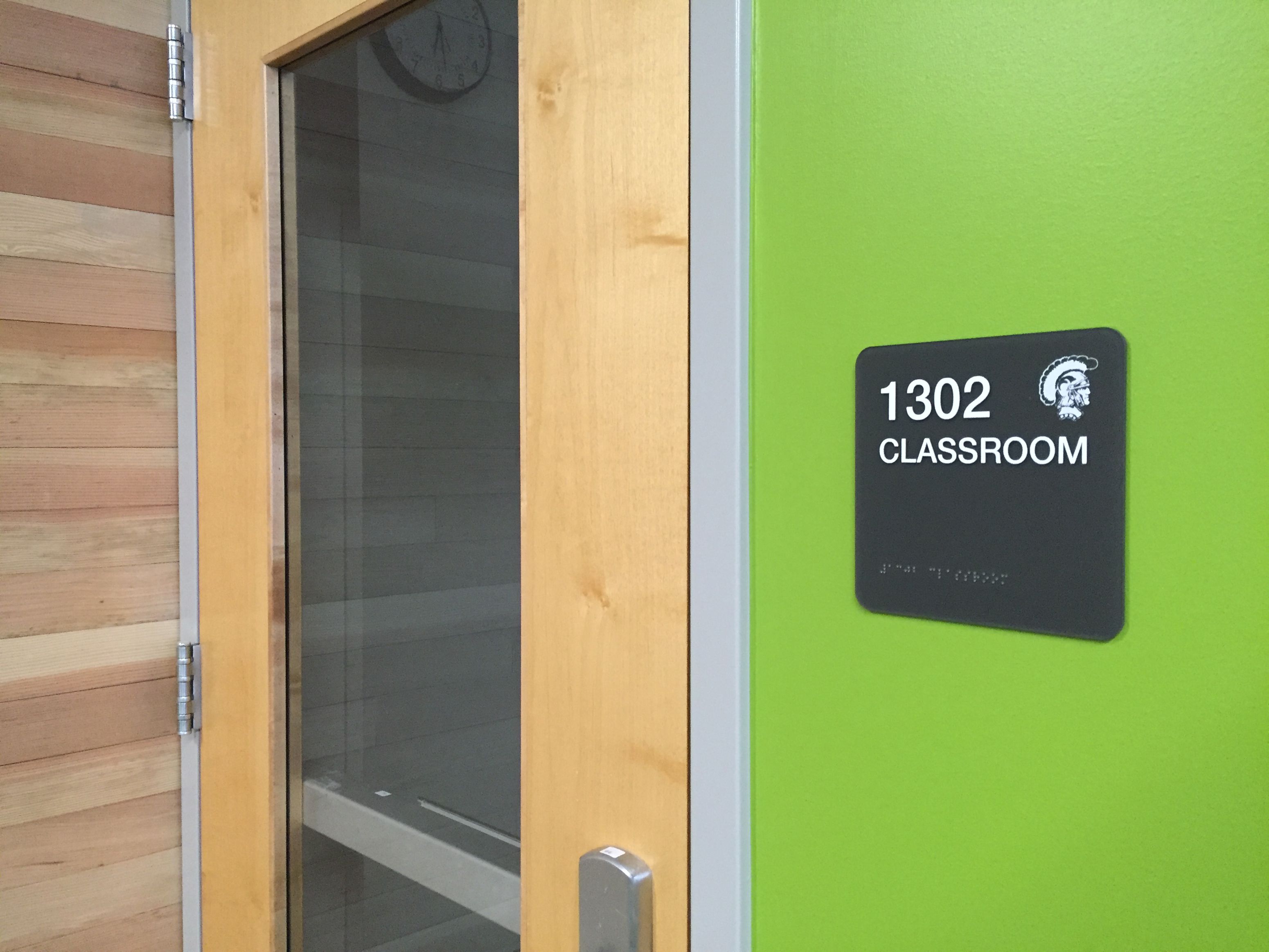 A green wall with a black sign displaying the room number "1320" in white text, above the word "CLASSROOM." The sign also includes a small image of a gladiator helmet and has Braille text at the bottom.
