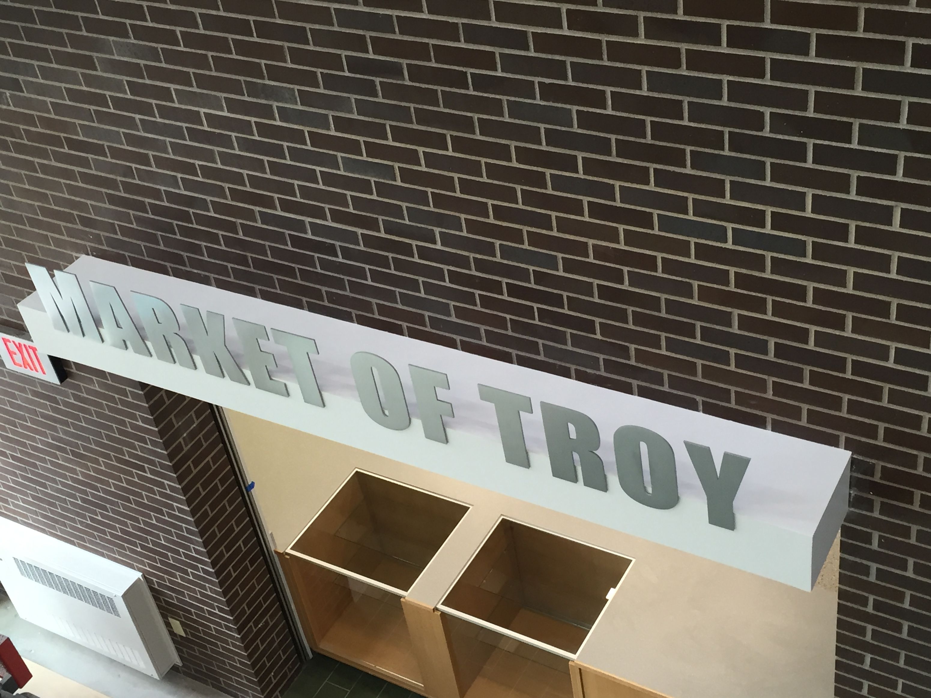 A white rectangular sign with gray 3D letters reads "MARKET OF TROY" mounted above a doorway. The surrounding walls are made of beige brick. Below the sign are empty display cases and a section of tiled floor is visible. An exit sign is visible to the left.