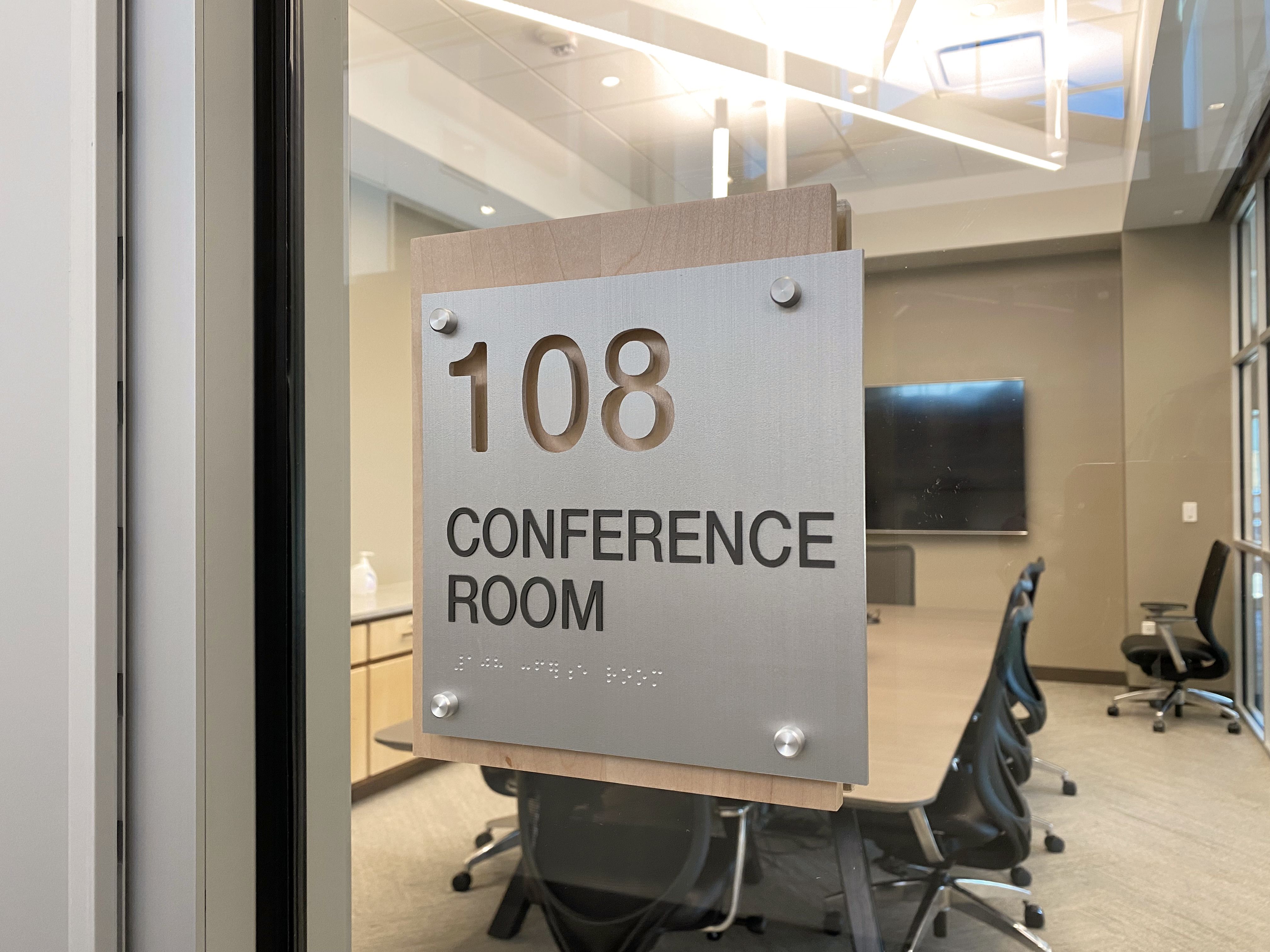 A glass door with a silver sign that reads "108 Conference Room" in black letters. Inside the room, a large table surrounded by several office chairs is visible. The room is well-lit and a large screen is mounted on the wall in the background.