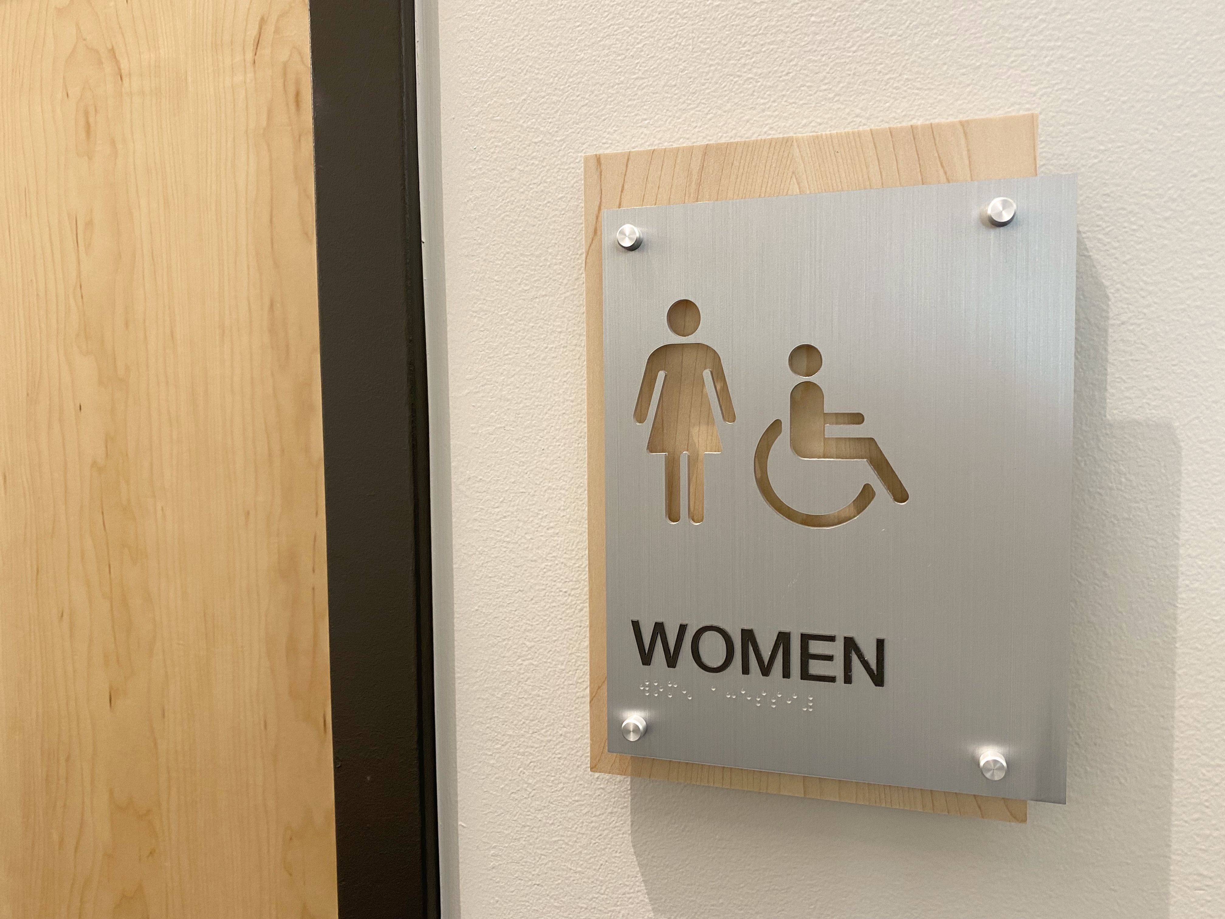 A sign on a wall indicates a women's restroom and features symbols for both women and accessibility. The sign is made of a metallic material with "WOMEN" written below the symbols. The door to the restroom is partially visible on the left.
