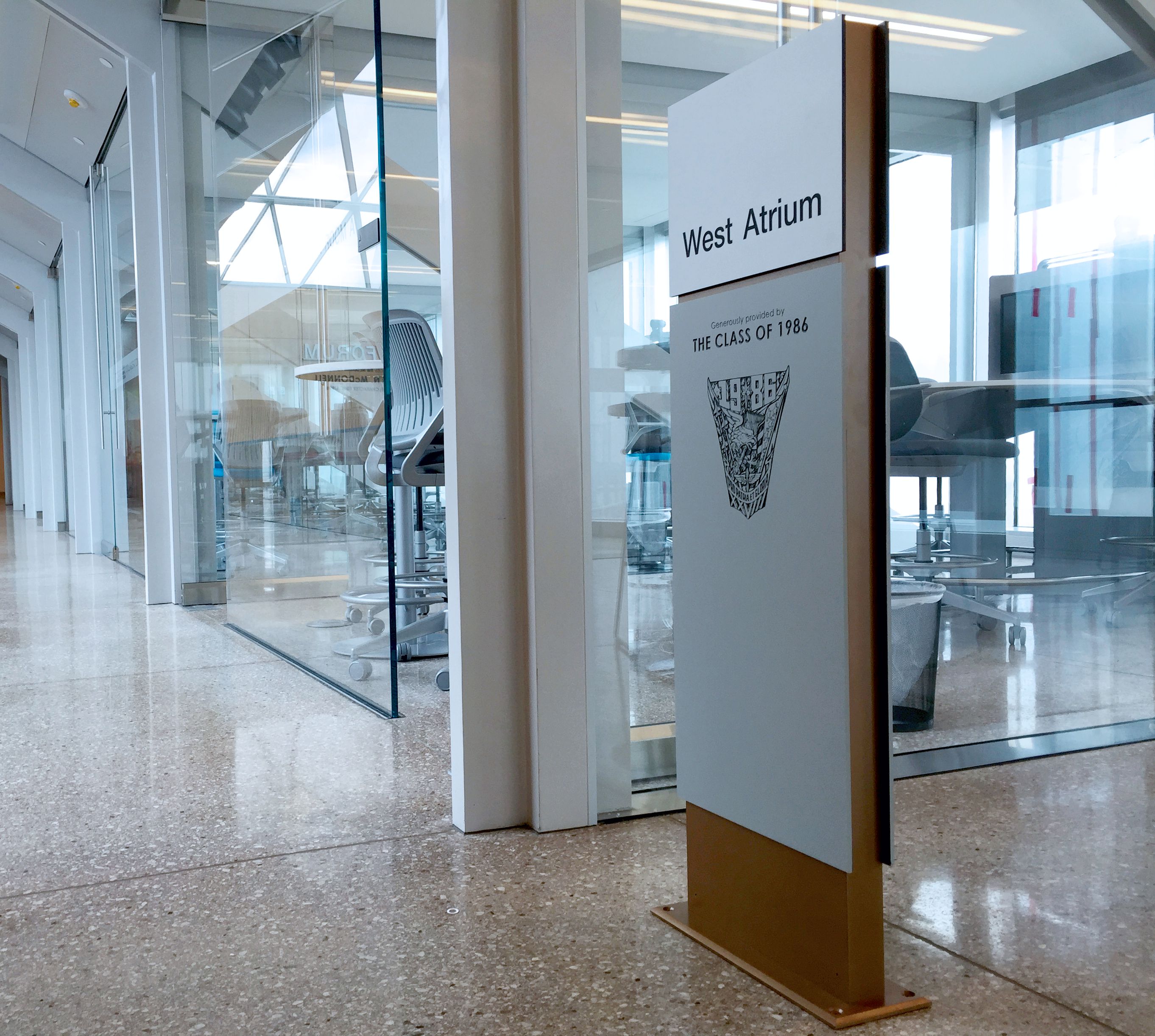 A modern indoor hallway with polished floors leads to a glass-walled room on the right, labeled "West Atrium." A sign commemorates "The Class of 1986," featuring an intricate design. Glass doors and large windows allow natural light to brighten the corridor.