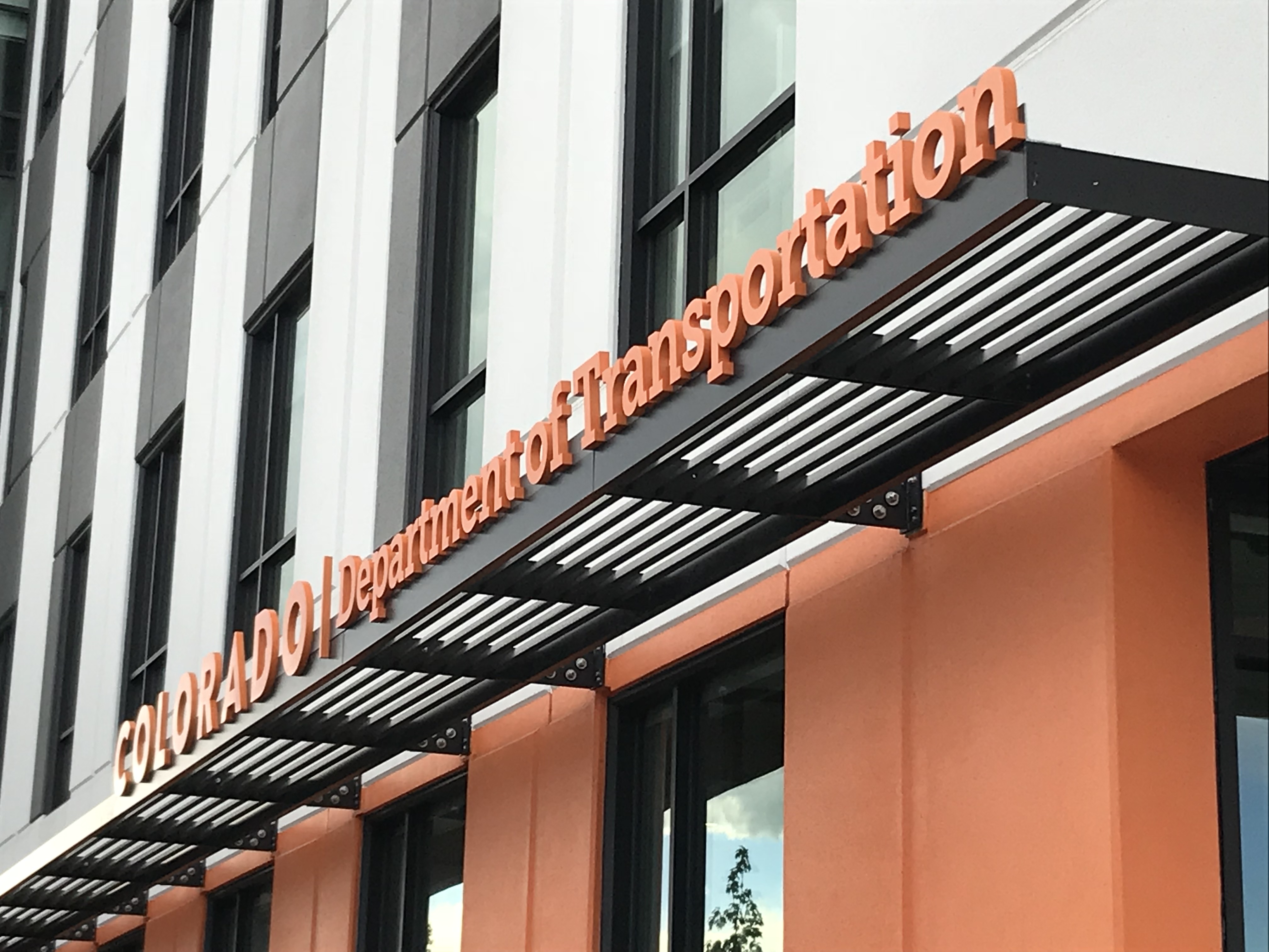 A modern building with the sign "Colorado Department of Transportation" displayed in orange letters above the entrance. The facade features a combination of white and orange panels with large windows.