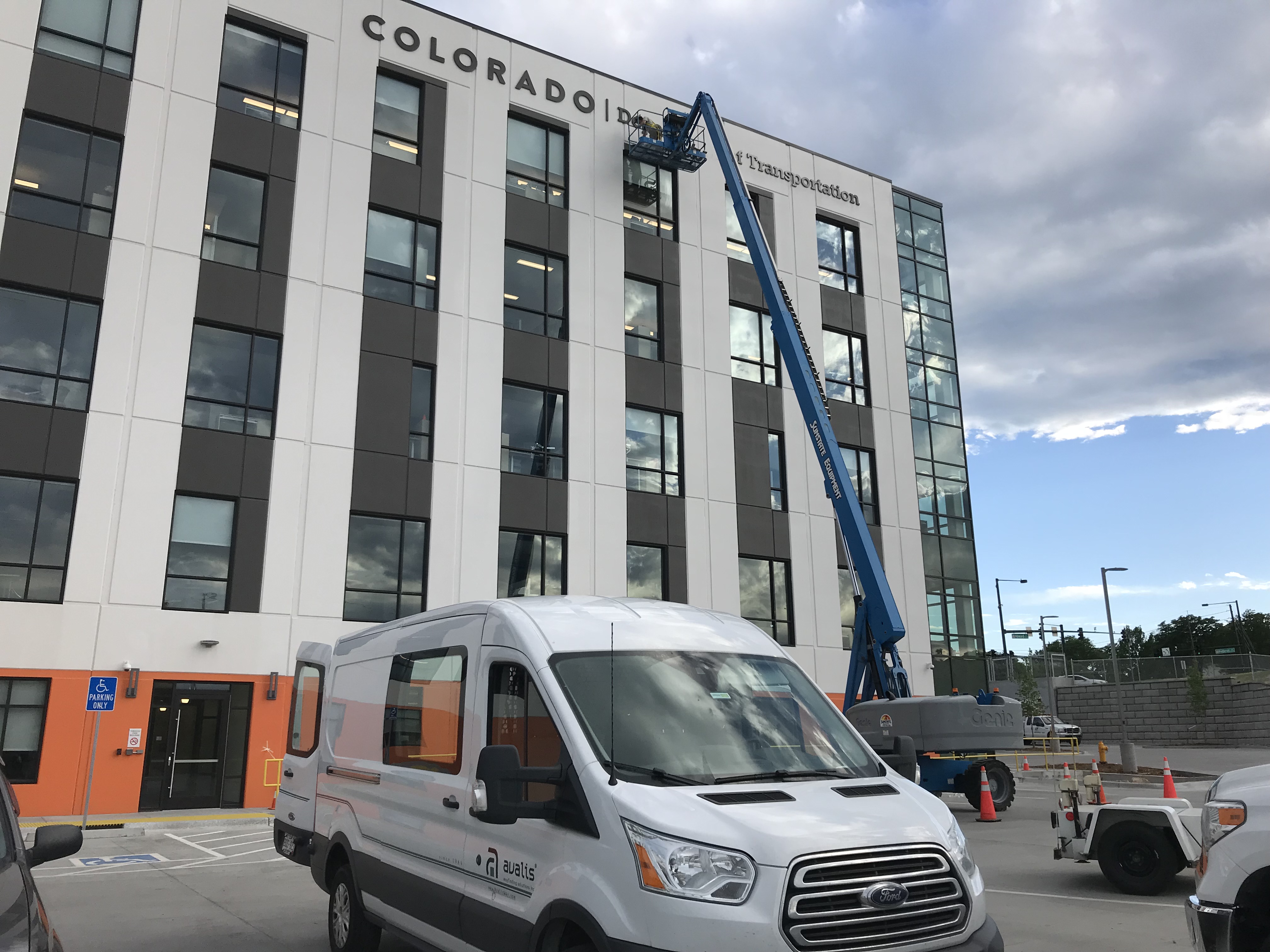 A worker is elevated in a blue boom lift near the windows of a multi-story building labeled "Colorado Department of Transportation." Orange cones surround the lift.