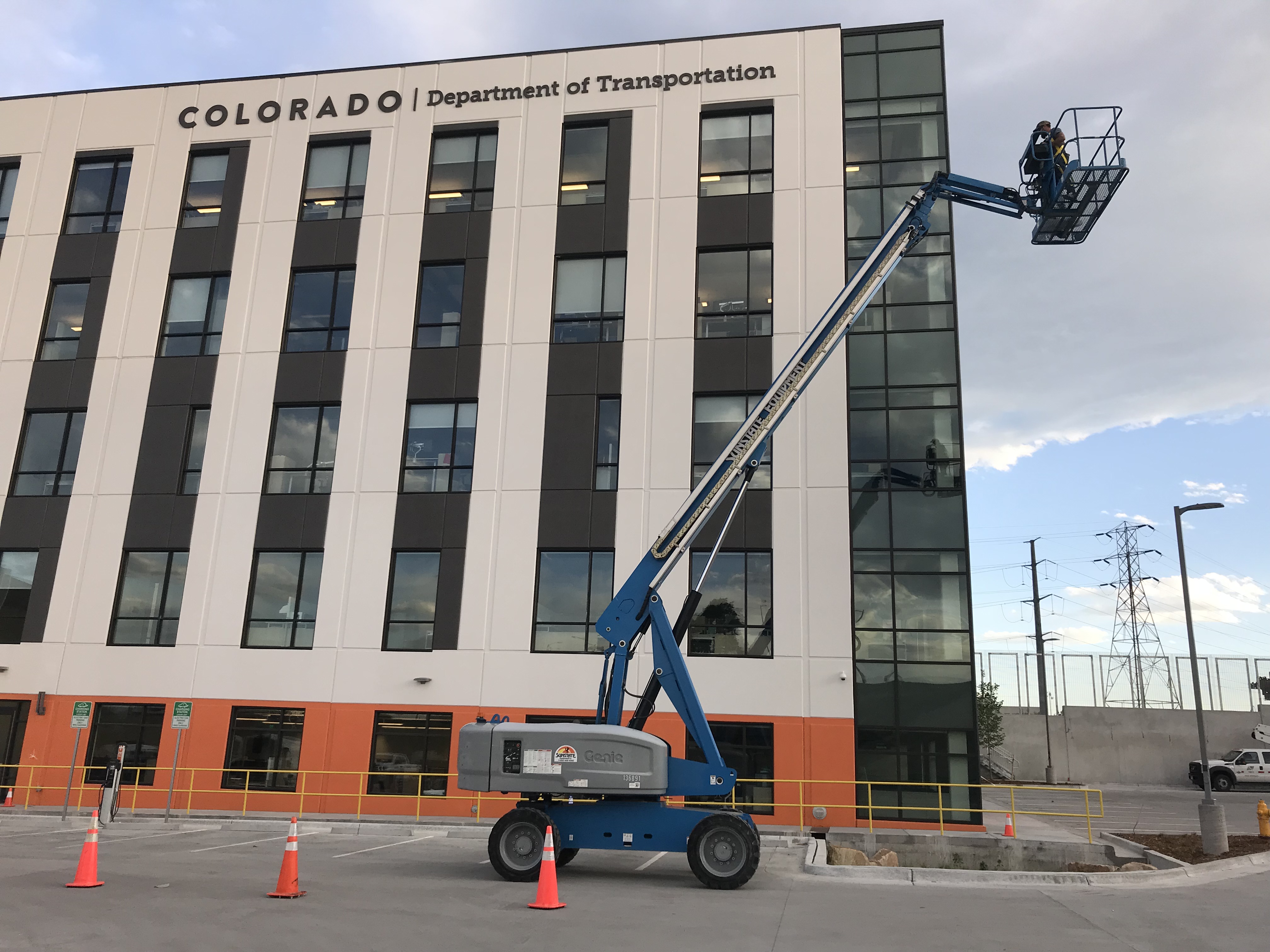 A worker is elevated in a blue boom lift near the windows of a multi-story building labeled "Colorado Department of Transportation." Orange cones surround the lift.