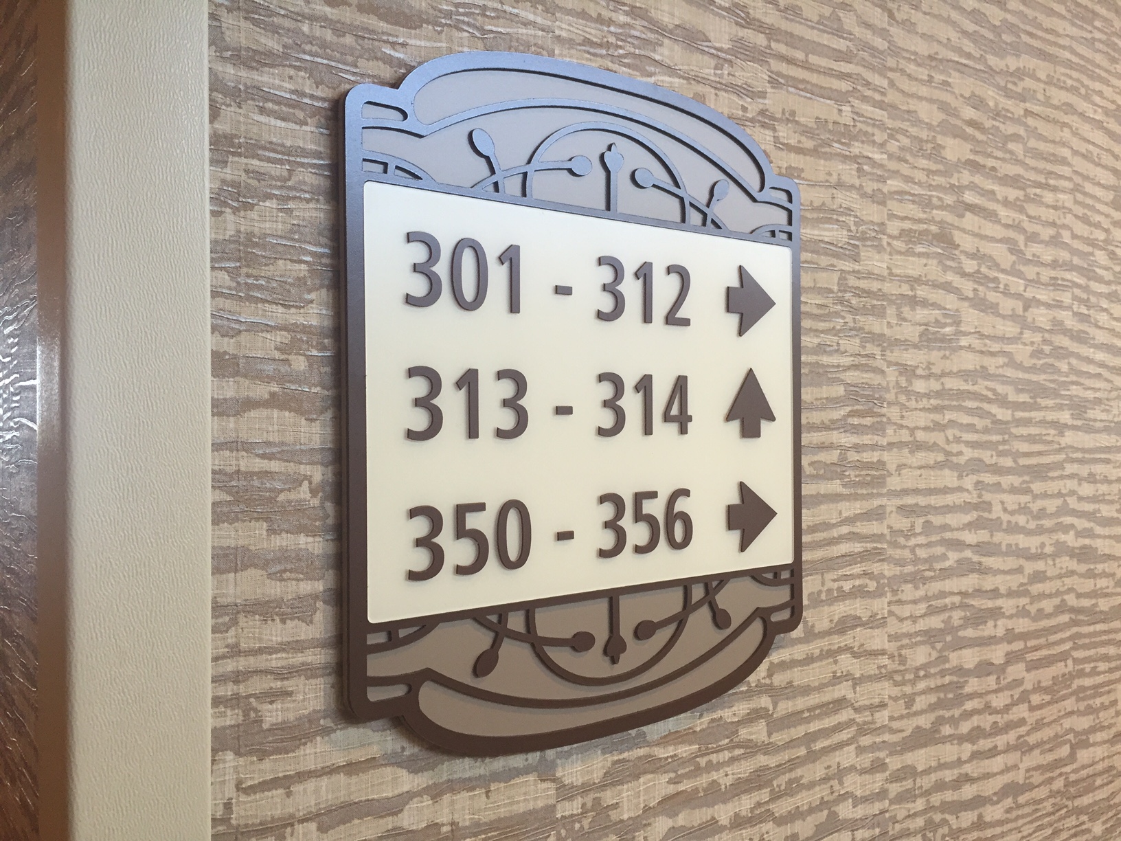 A wall-mounted sign in a hallway displays room numbers with arrows indicating directions. Rooms 301-312 are to the right, rooms 313-314 are straight ahead, and rooms 350-356 are to the left. The sign has a decorative border and is mounted on a textured wall.