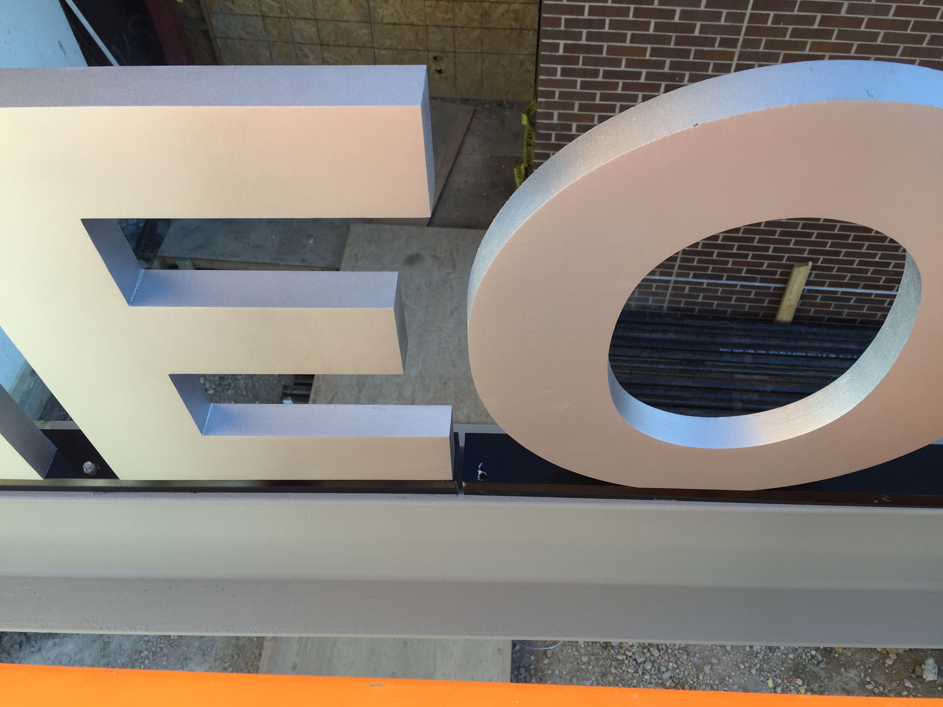 Close-up view of large metallic letters "E" and "O" on a sign, partially showing other letters. The background includes a brick wall and construction materials on the ground.