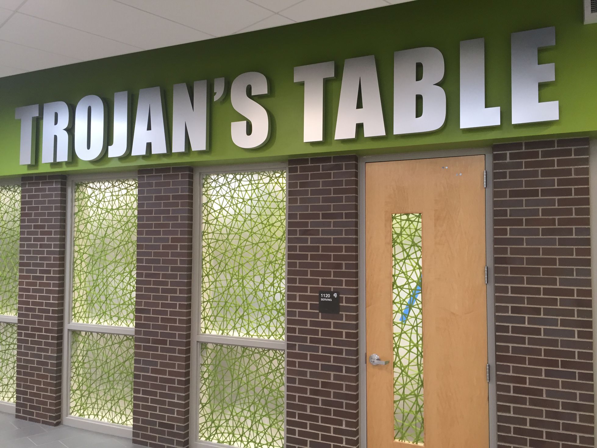 A dining area features a sign reading "TROJAN'S TABLE" in bold, green letters above frosted glass windows and a wooden door. 