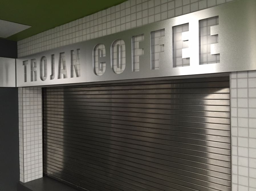A closed metal security shutter with "TROJAN COFFEE" in large, cut-out block letters above it. The background features white tiled walls, and the sign is illuminated from above.